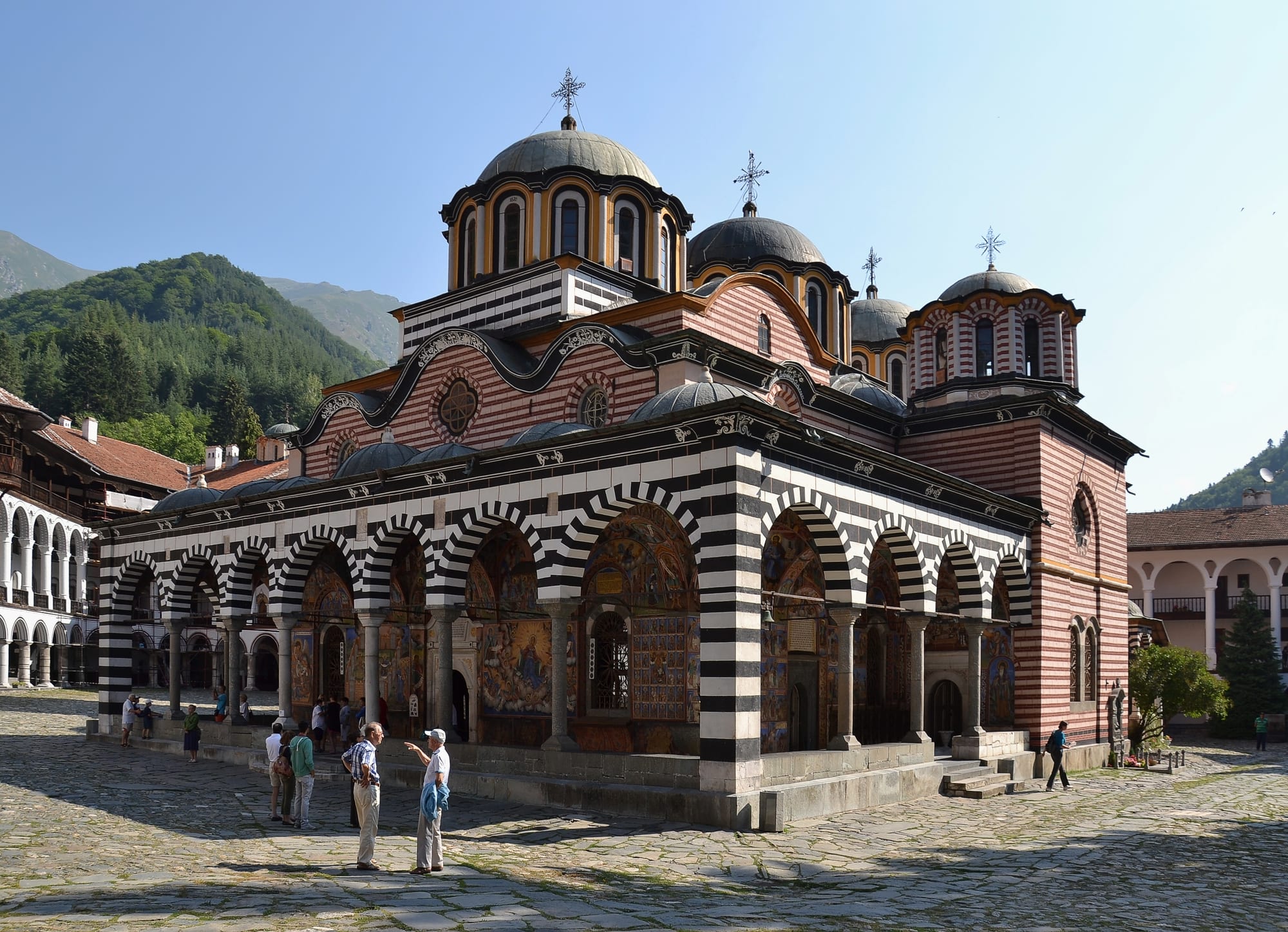 A large monastery courtyard with striped arcaded galleries around a domed church, backed by forested mountains