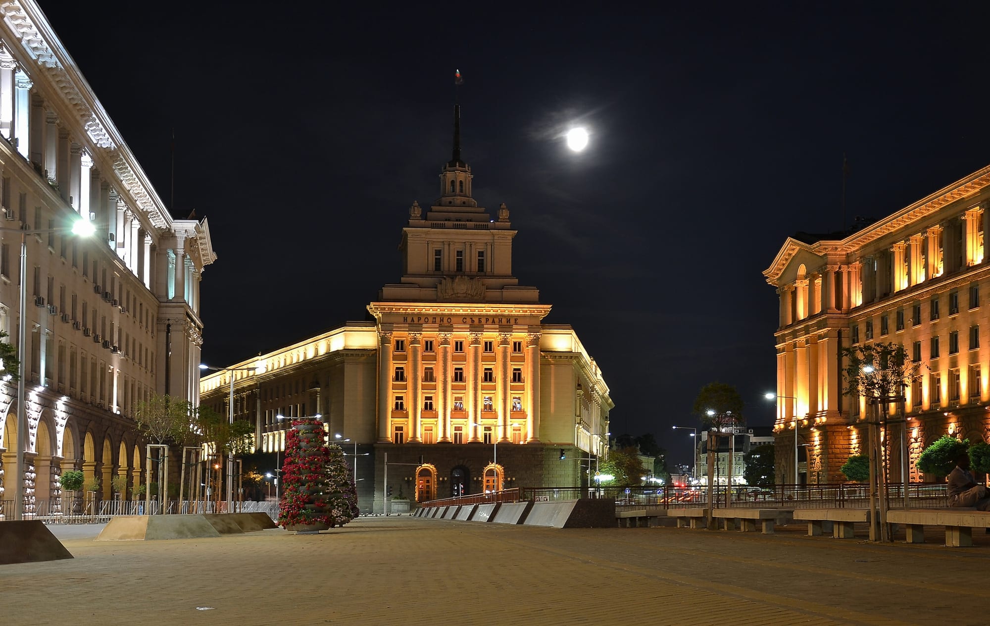A floodlit Stalinist-style building with a colonnaded tower and spire at the end of a paved square at night, under a full moon