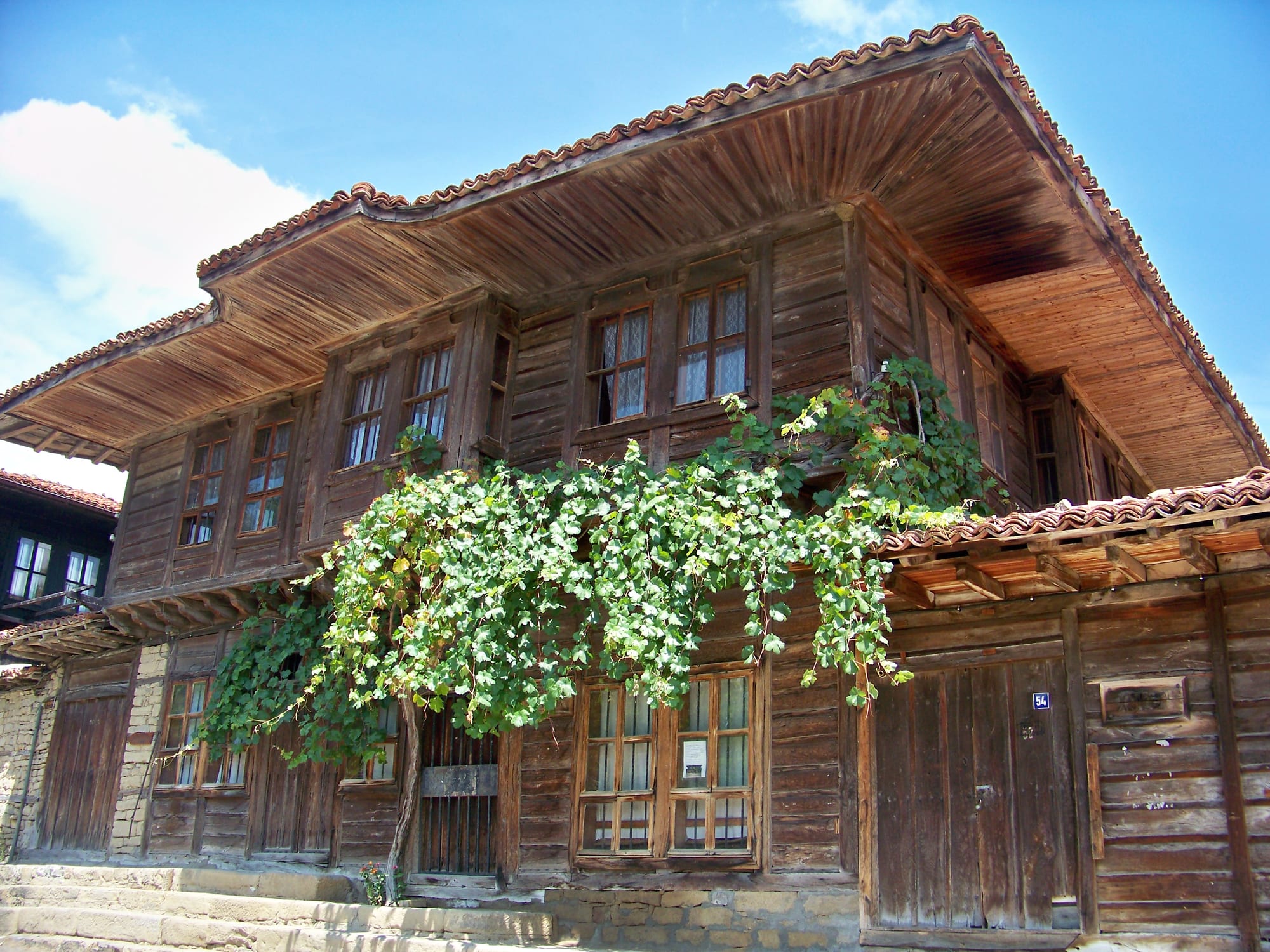 A traditional two-storey wooden Bulgarian house with a tiled roof and a grapevine trailing across its facade