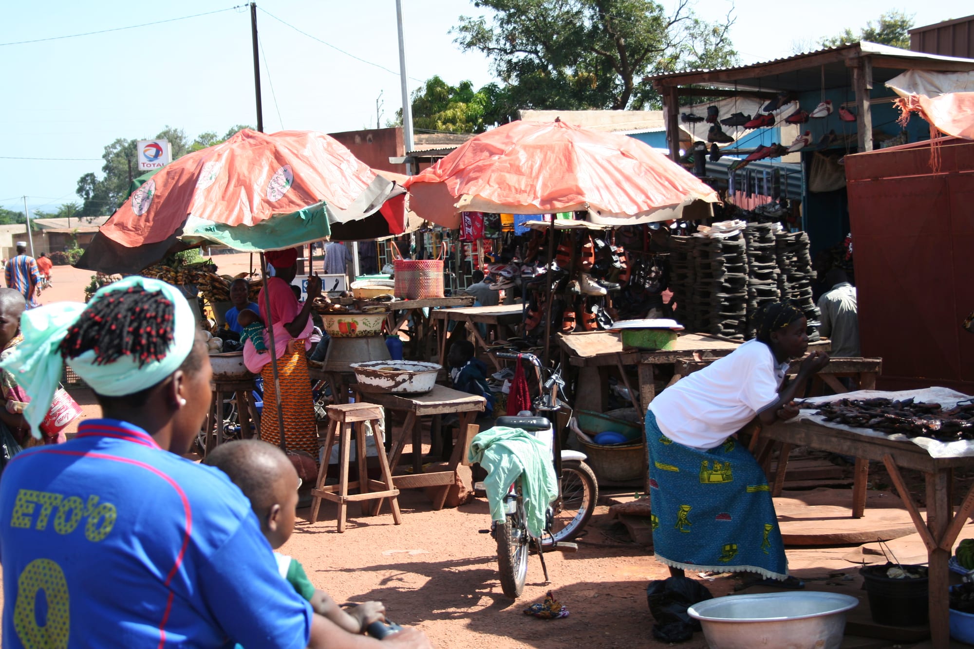 Vendors selling shoes and other goods under large market umbrellas in Banfora, with a bicycle parked beside a stall and shoppers moving between tables