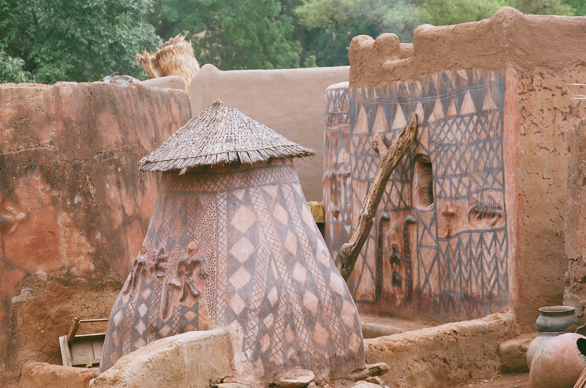Earthen buildings covered in painted geometric patterns of diamonds, triangles and zigzags in red, black and white, including a conical building with a thatched roof and figures moulded in relief, with clay pots in the foreground