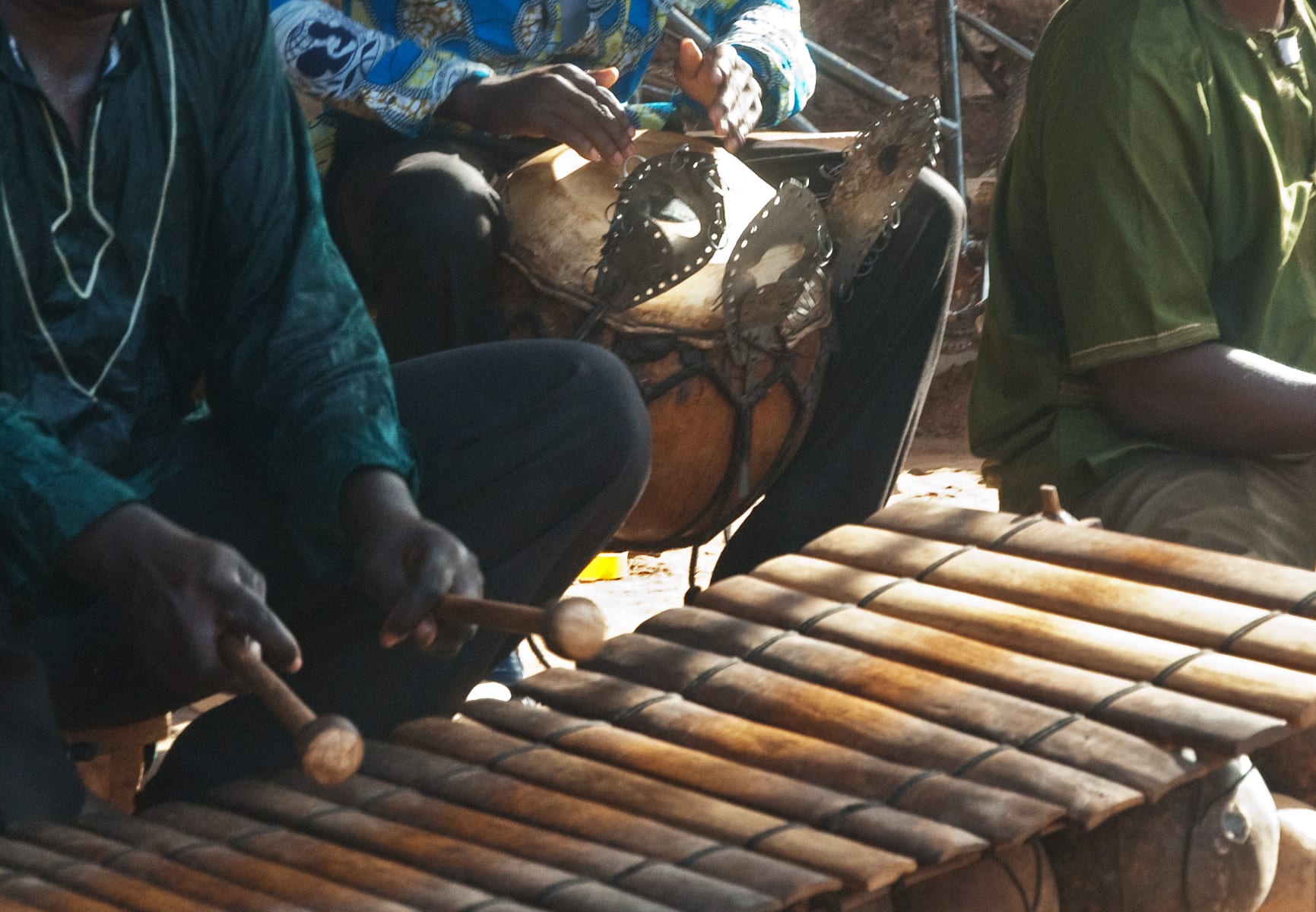 Close view of a musician's hands striking the wooden keys of a balafon with two beaters, while another musician behind plays a large gourd drum fitted with metal rattles