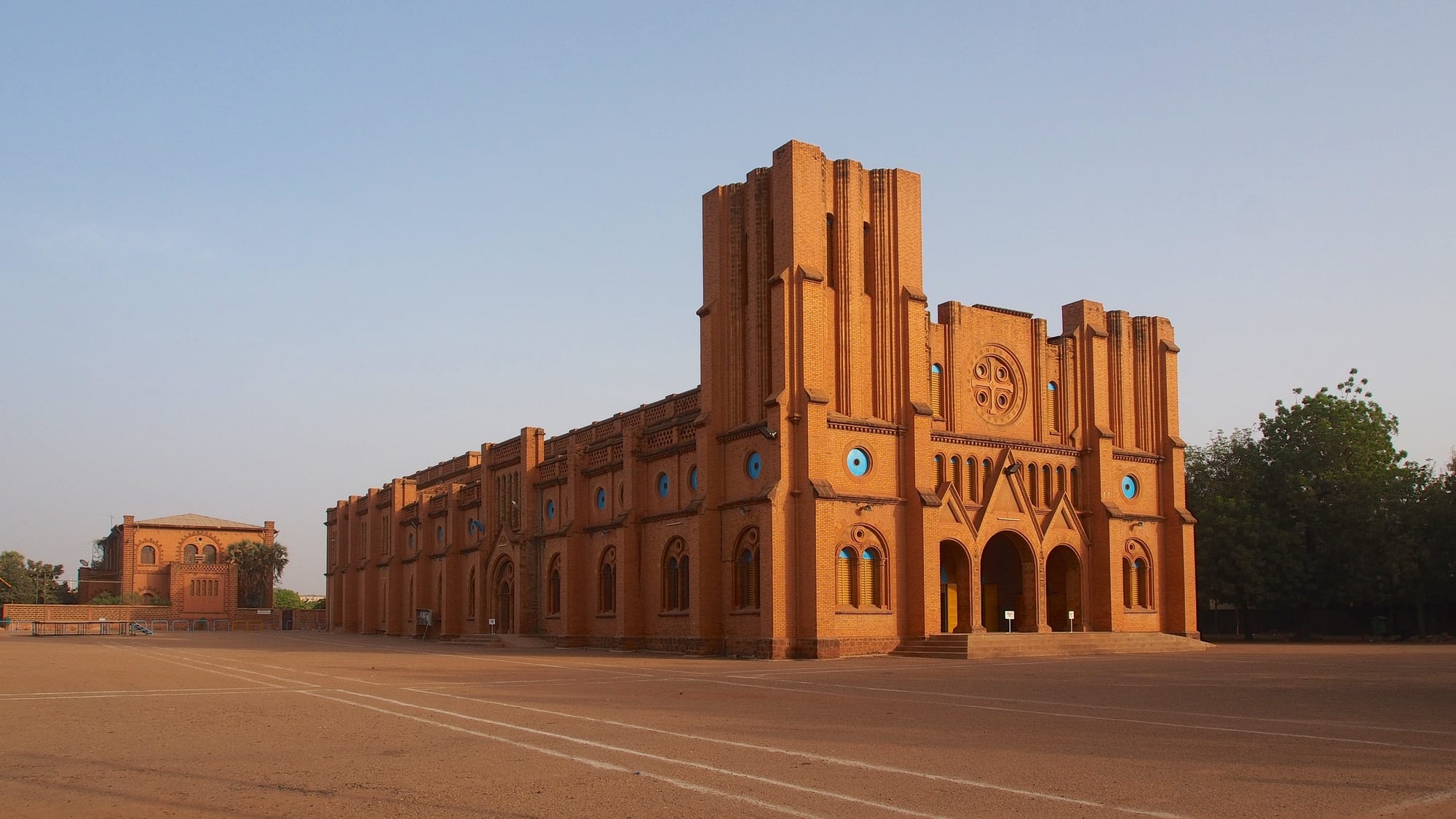 A large red-brick cathedral with a tall square tower, round blue windows and arched doorways, standing on a wide open square of bare earth under a clear sky