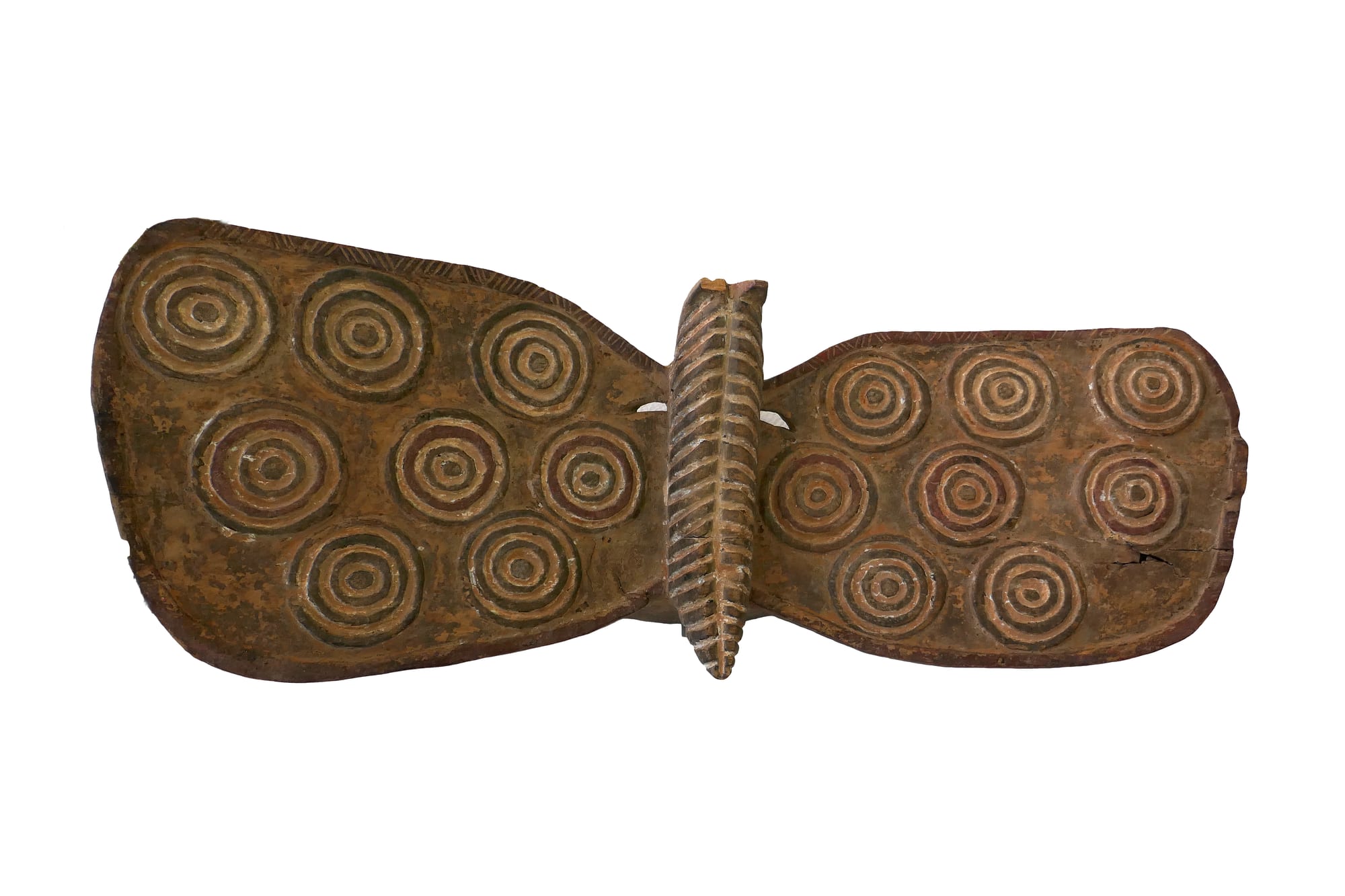 A wide, flat wooden mask shaped like a butterfly, its two wings carved with rows of concentric circles in brown, red and cream, with a ridged central projection