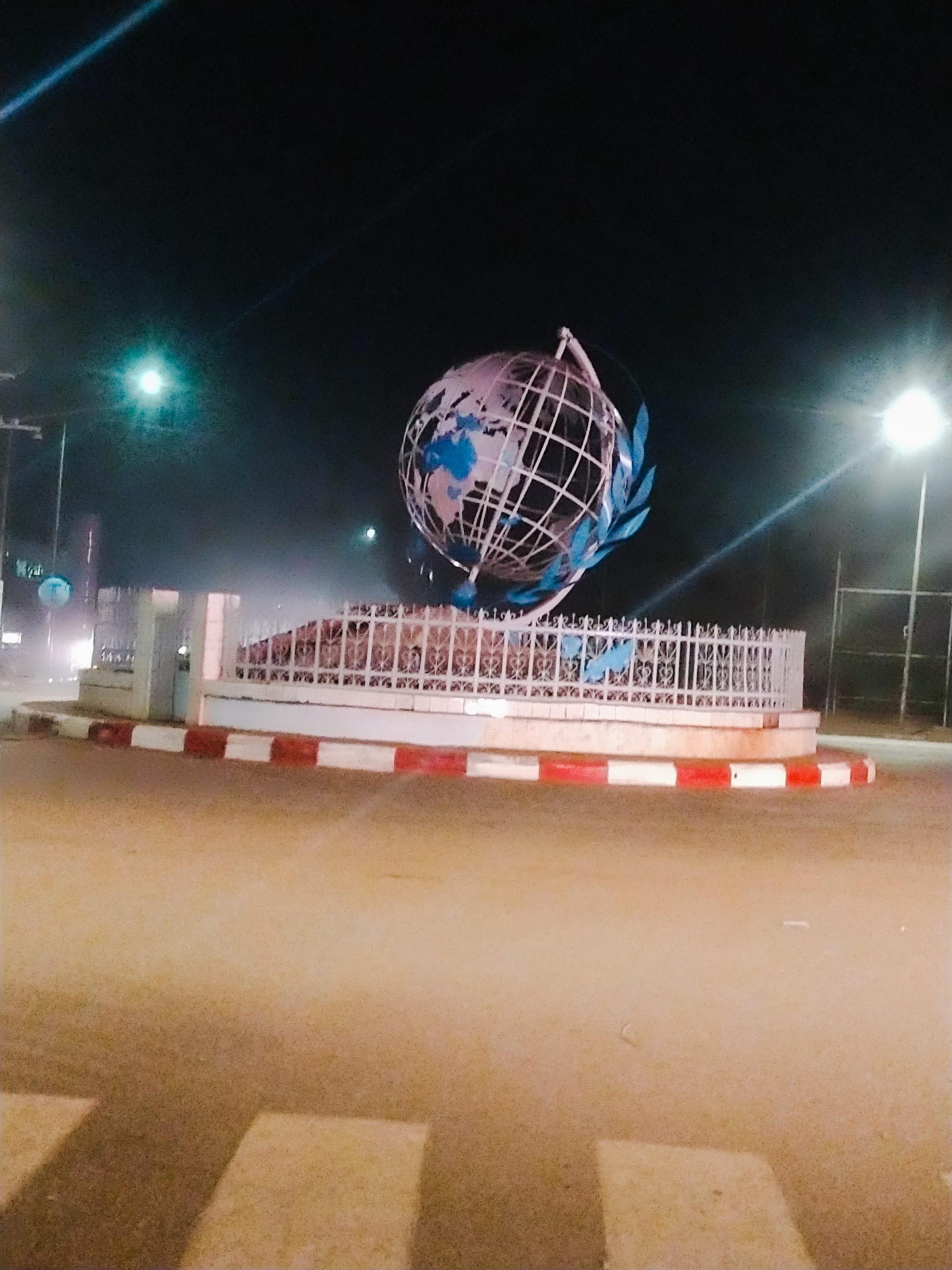 A busy city roundabout with a monument, motorbikes and low buildings under a hazy Sahel sky