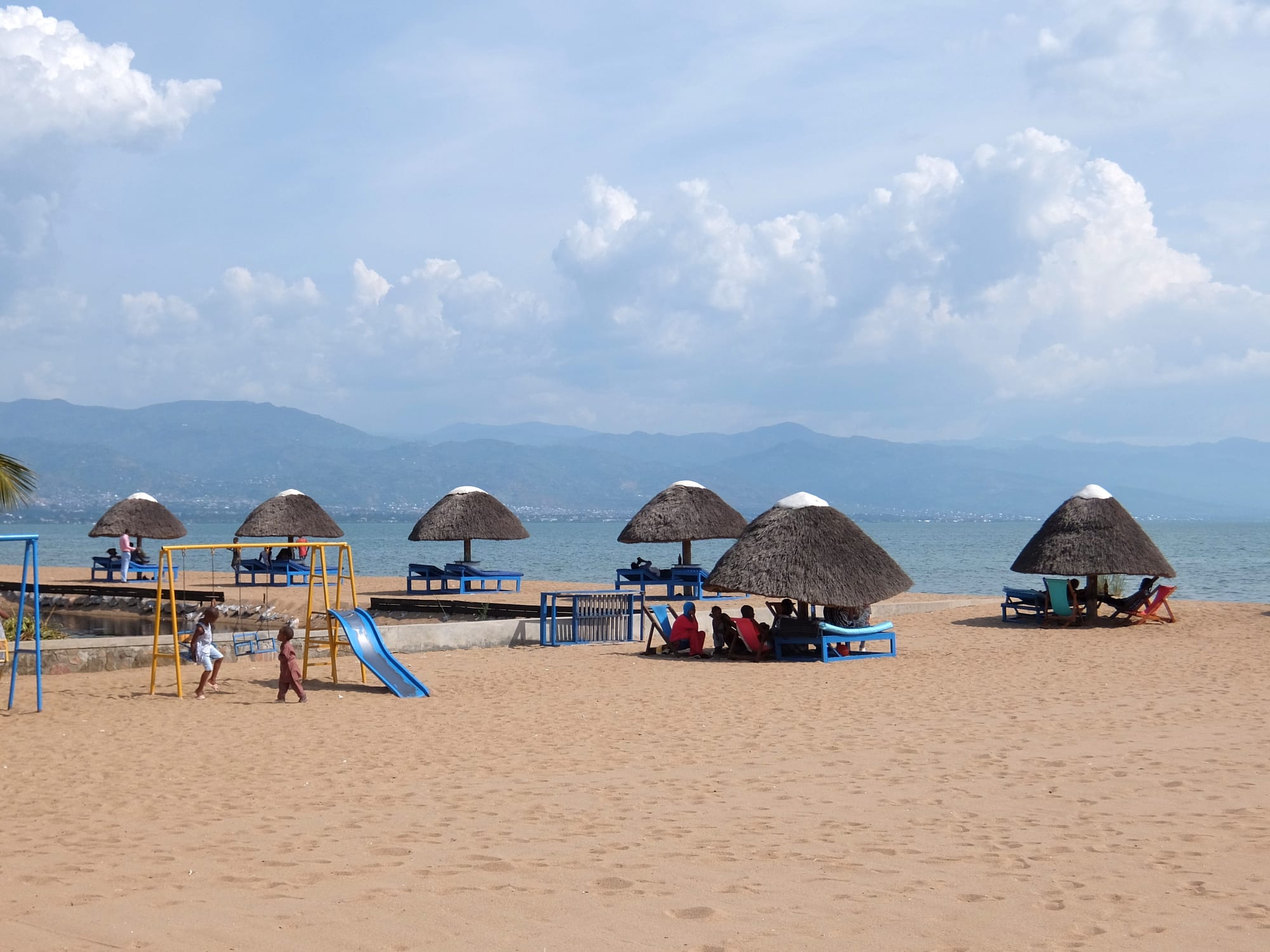 A sandy lake beach with palm-thatch shades and calm water, hills across the lake