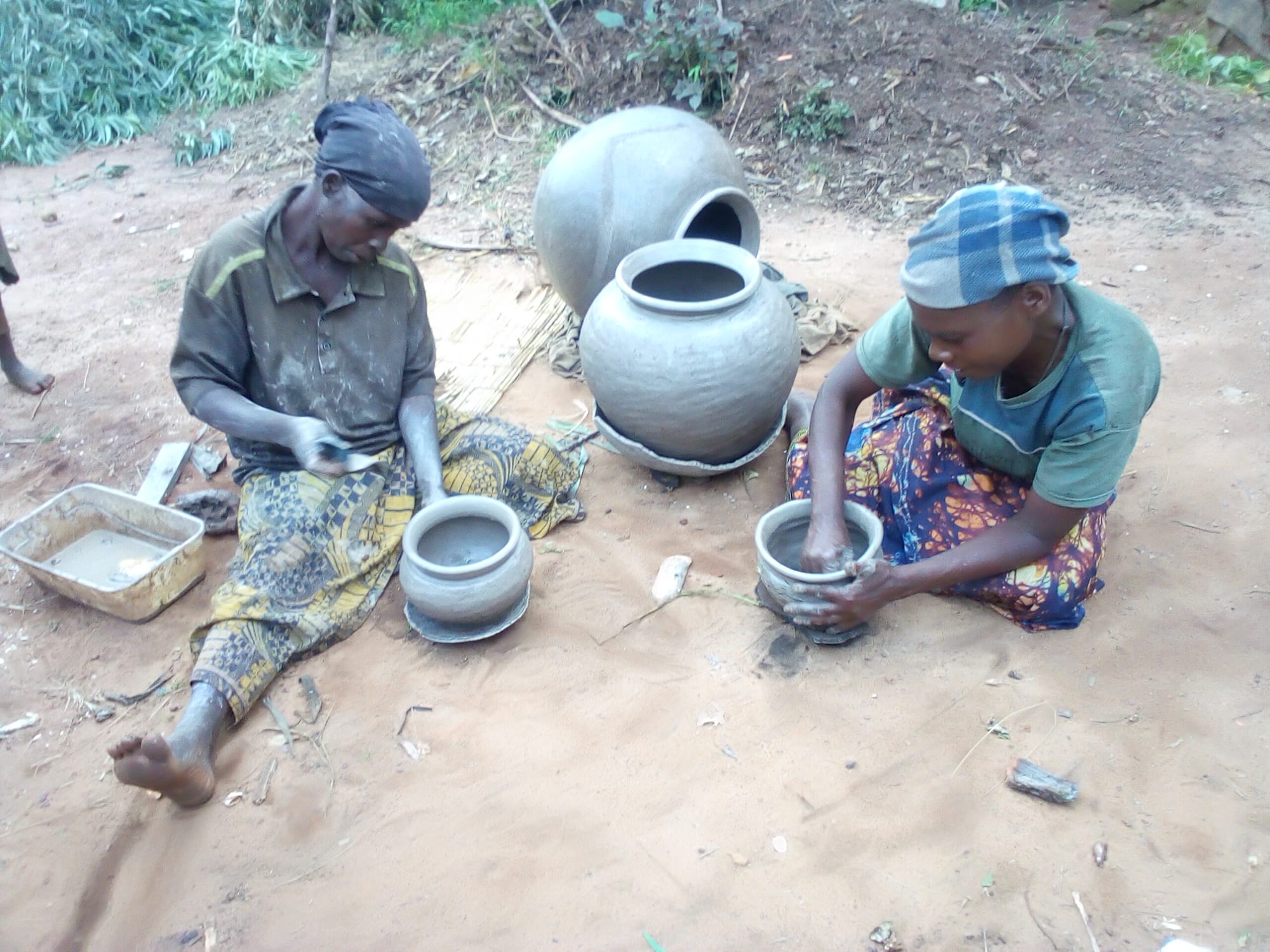 Two women sit on sandy ground shaping clay pots by hand, with two larger finished grey pots and a tray of clay beside them