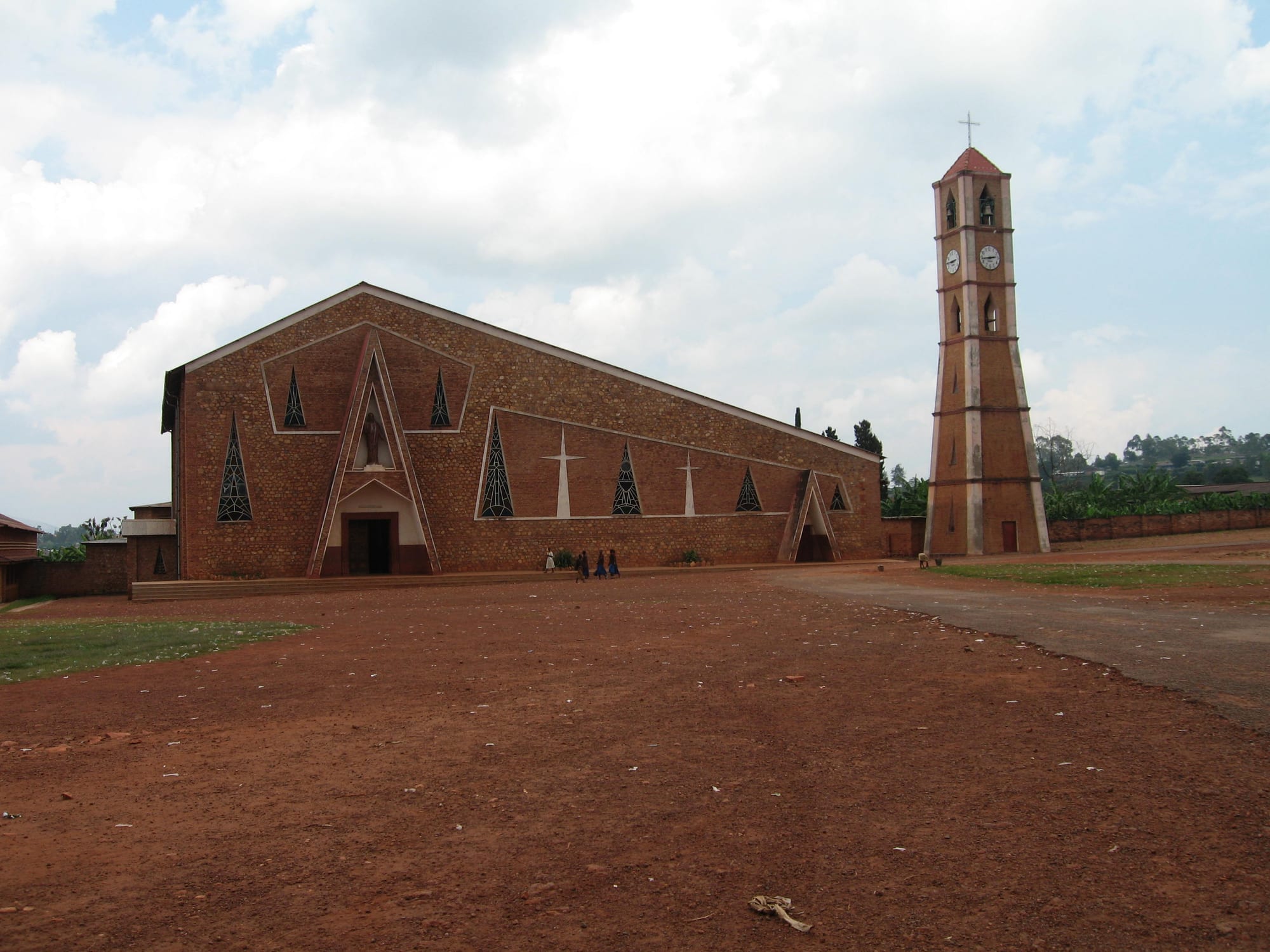 A large brick church with a pitched roof, triangular stained-glass windows and white crosses on its side wall, beside a tall freestanding brick bell tower with a clock, set on a wide red earth square