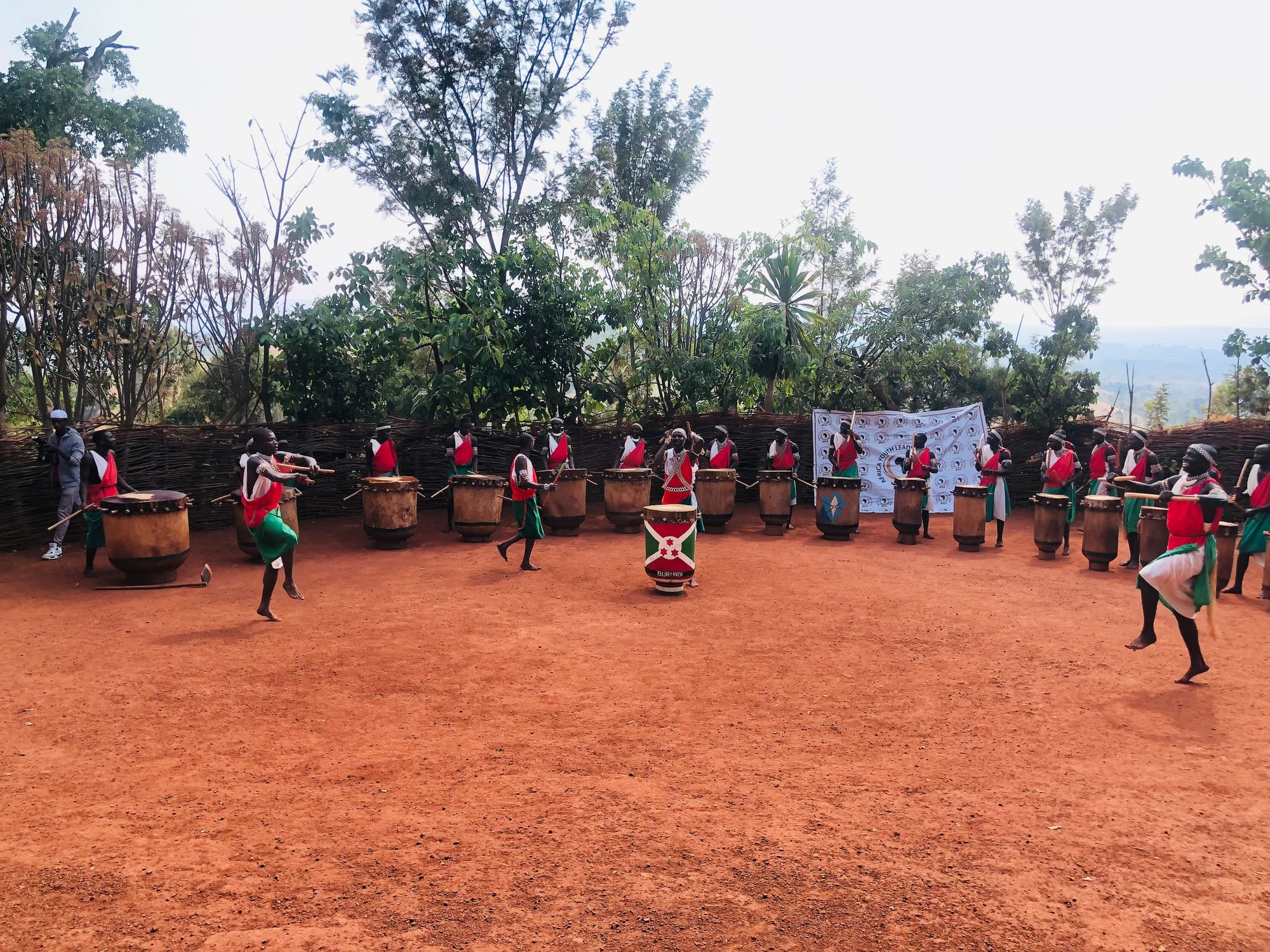 Drummers dressed in red, white and green stand behind a wide semicircle of tall wooden drums on a red earth ground, while dancers leap in front of them and a drum painted with the national flag stands at the centre