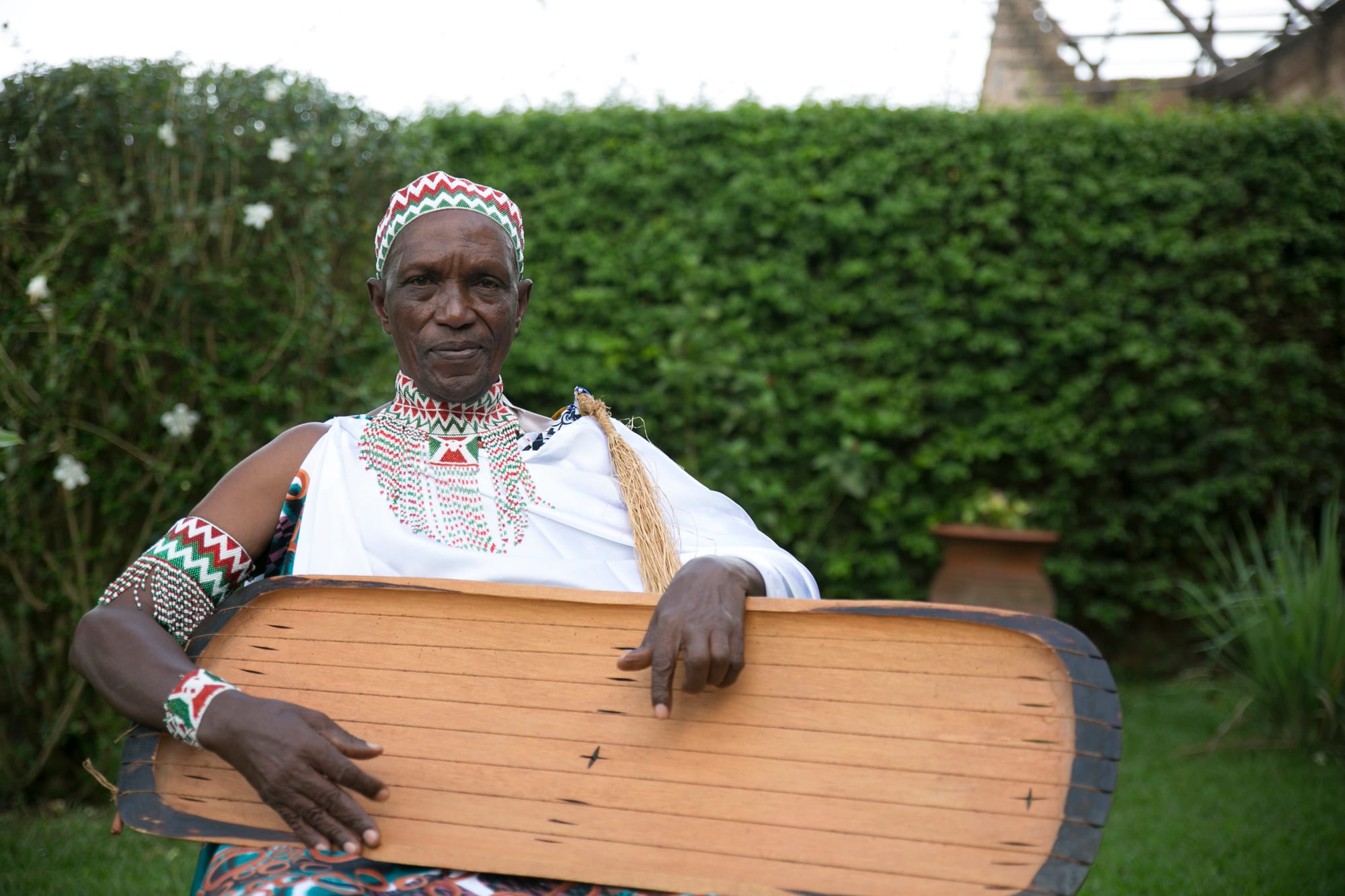 An elderly man in a white robe, a beaded collar and a patterned cap sits in a garden holding a long, shallow wooden trough zither across his lap