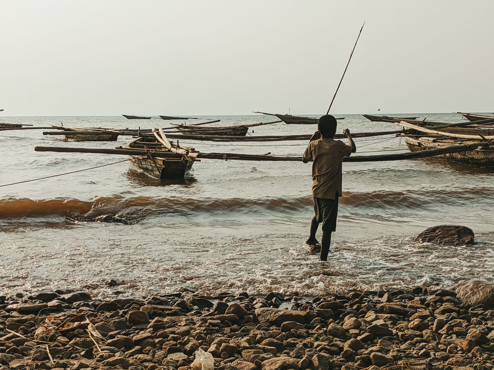 A wide calm lake meeting green hills under a hazy sky, with fishing boats near a sandy shore