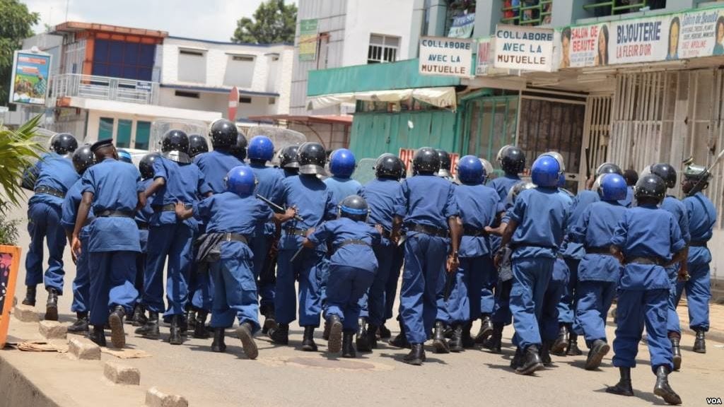 Riot police in helmets chasing fleeing protesters down a dirt street in a African city