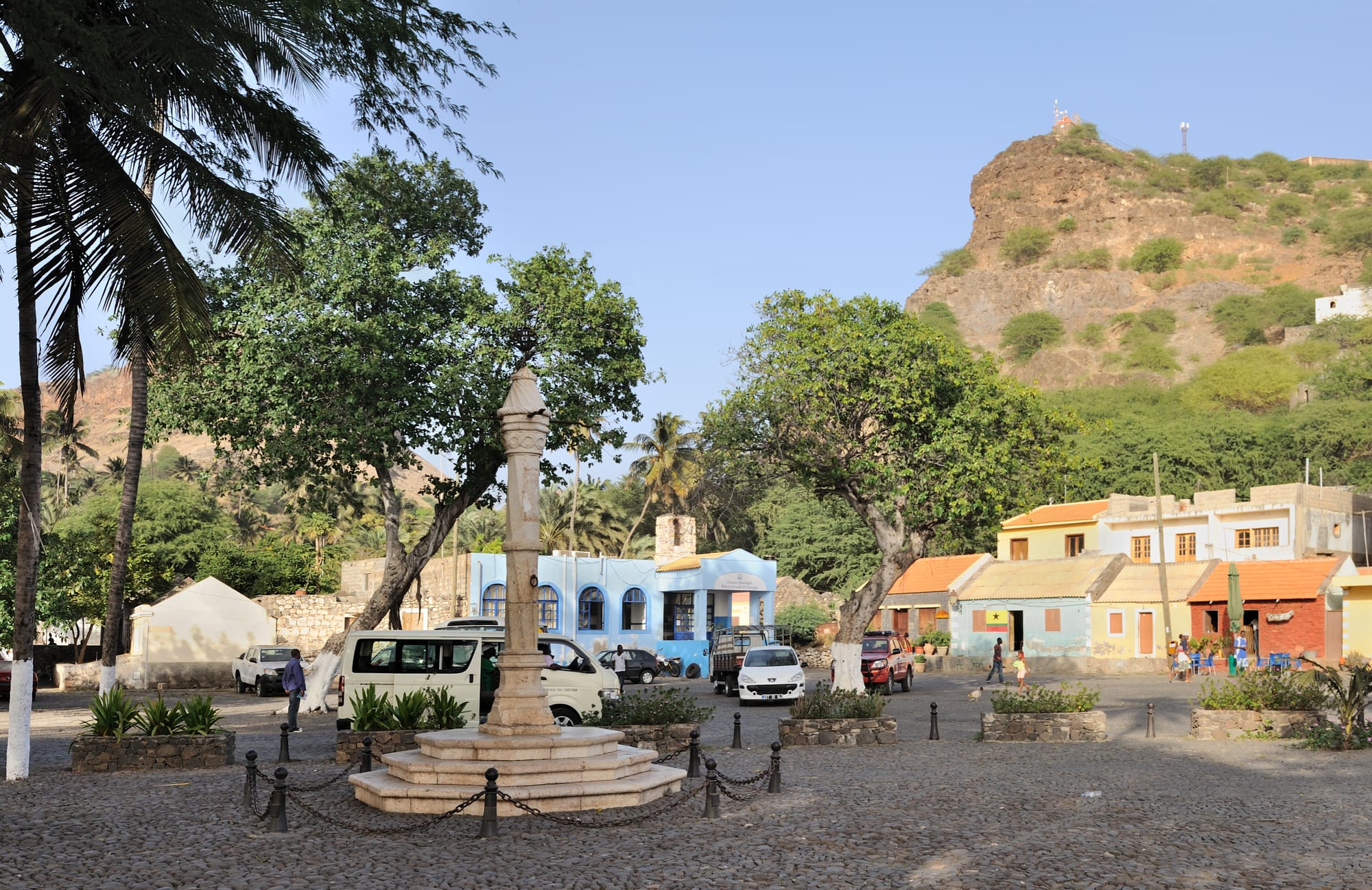 A stone pillory monument on a cobbled square in front of a whitewashed church, hills behind