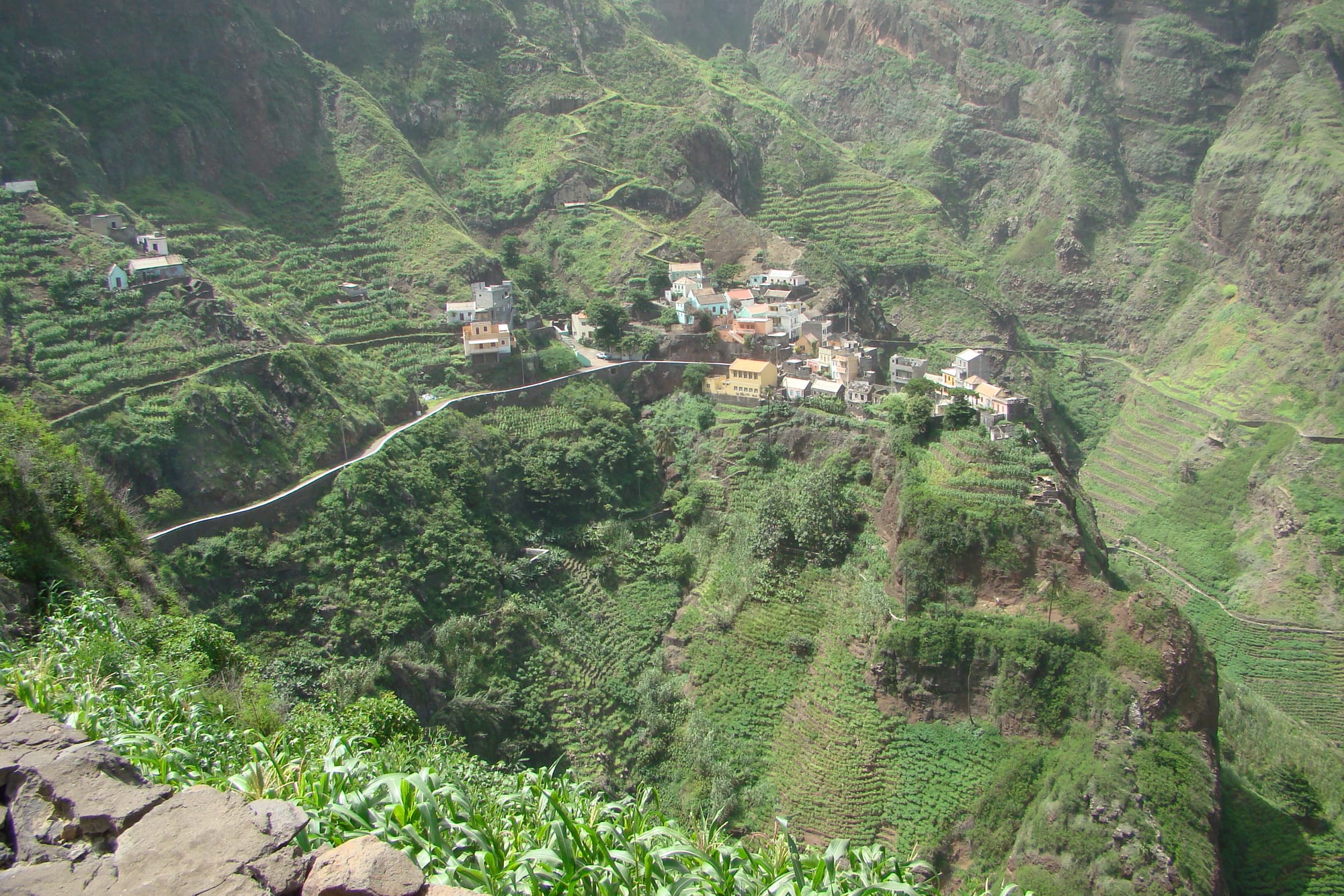A small village of pastel-coloured houses clustered on a narrow green terrace partway up a steep, deeply eroded mountain valley