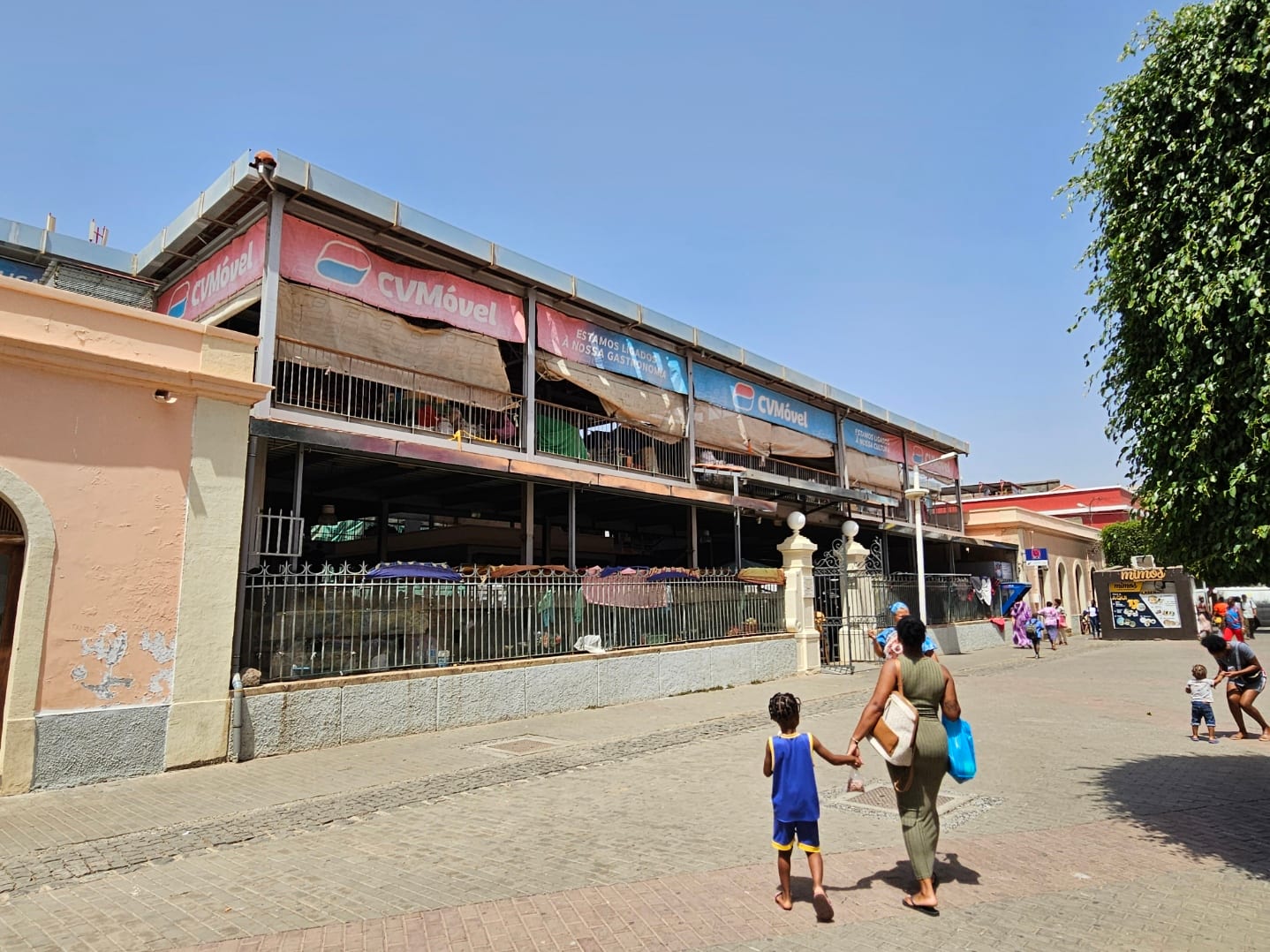 A woman and child walking hand in hand across a paved square past the covered municipal market in Praia, with other shoppers nearby
