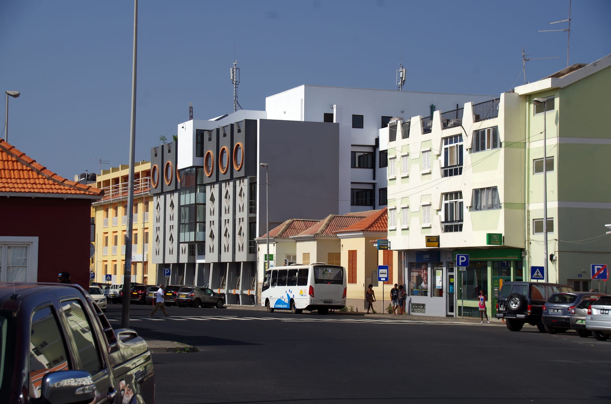 A city street in Praia with a white minibus, parked cars, low shopfronts and pedestrians crossing at a zebra crossing