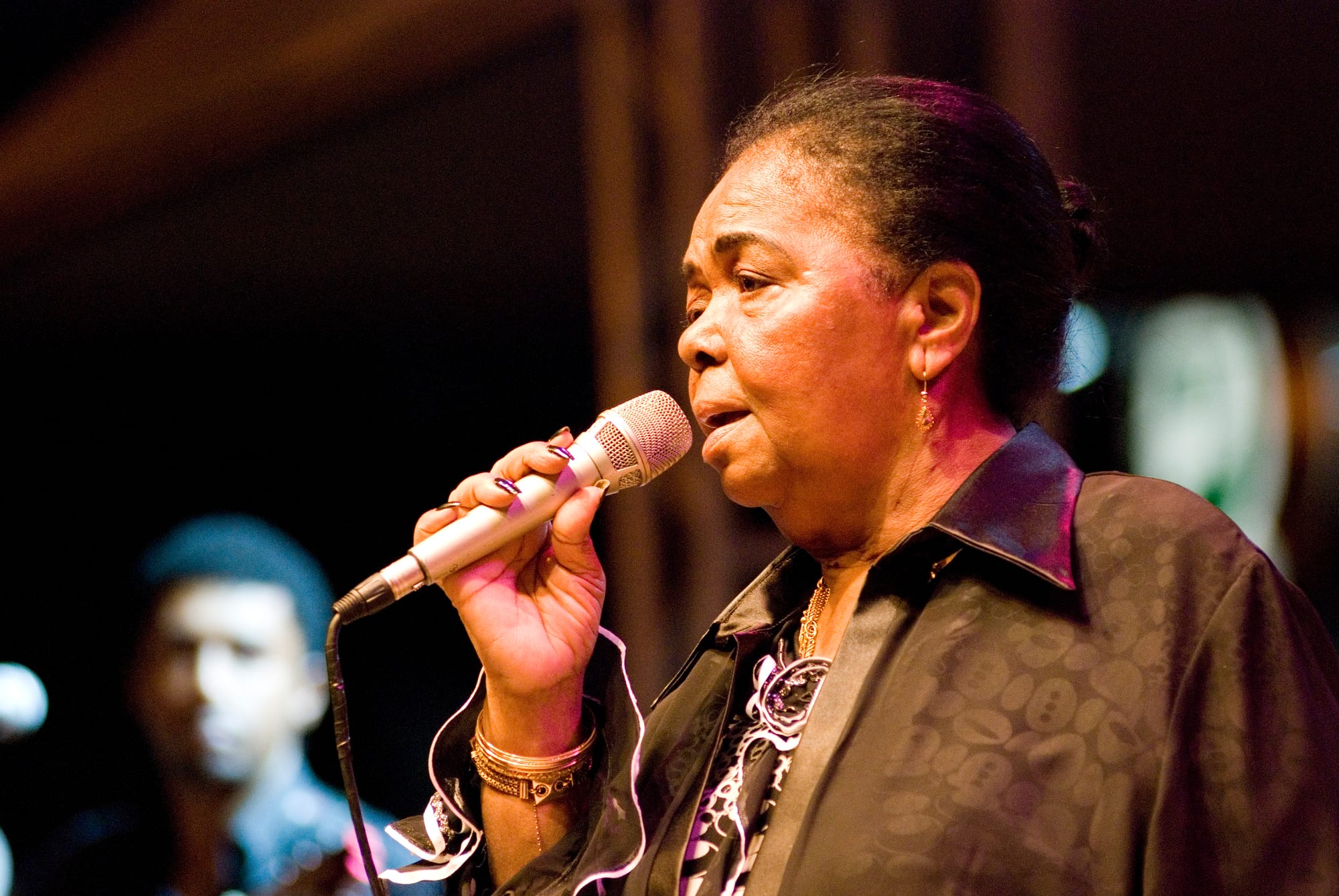 An older woman in a dark patterned blouse and gold bracelets sings into a microphone held close to her face, with a blurred musician behind her