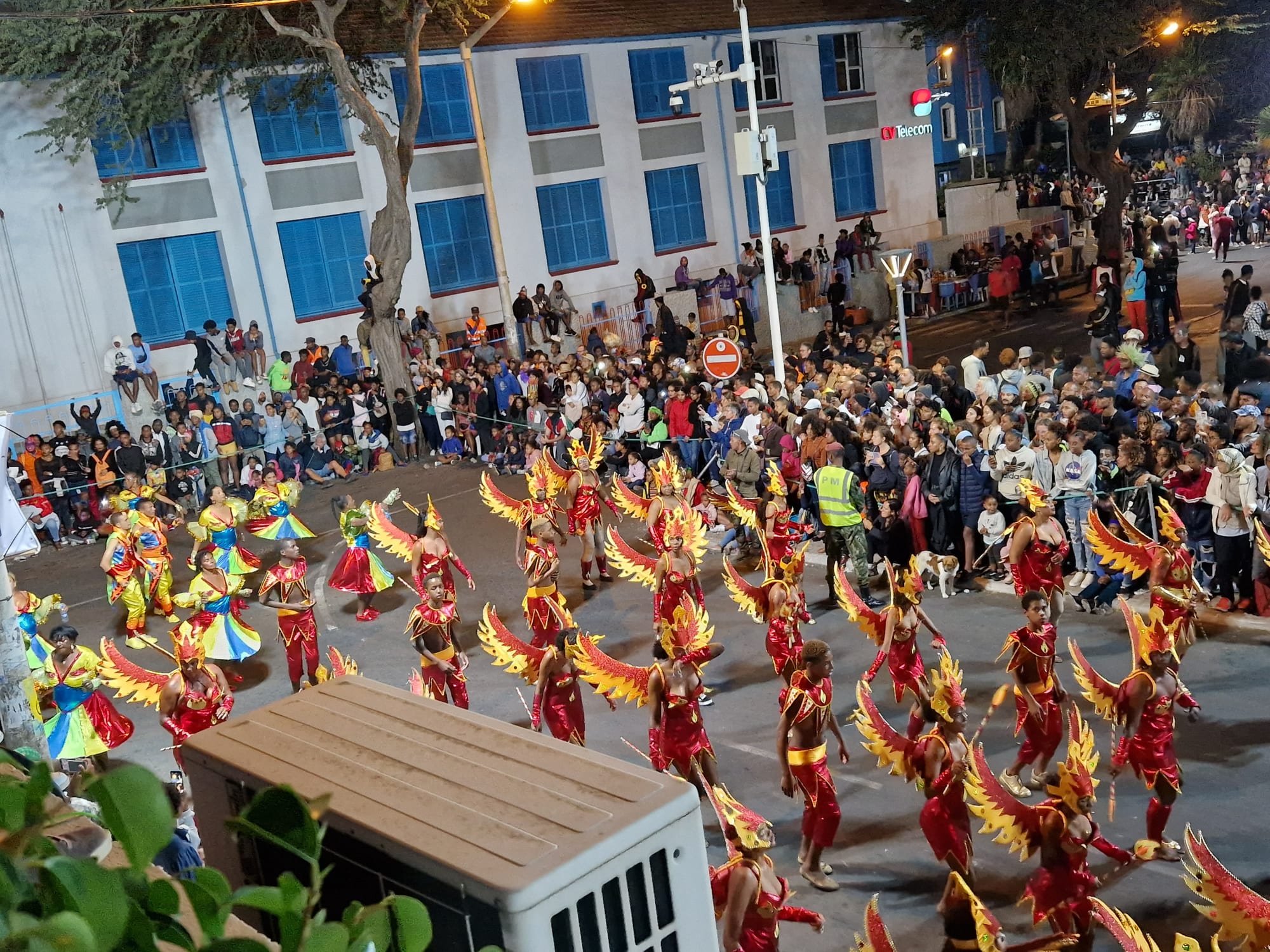 Rows of dancers in red and gold costumes with large feathered wings parade along a street at night, watched by dense crowds on the pavement and on the steps of a building with blue shutters