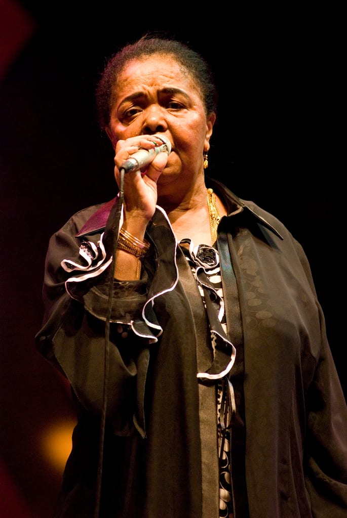 An older woman singing barefoot on stage, eyes closed, in a simple dress