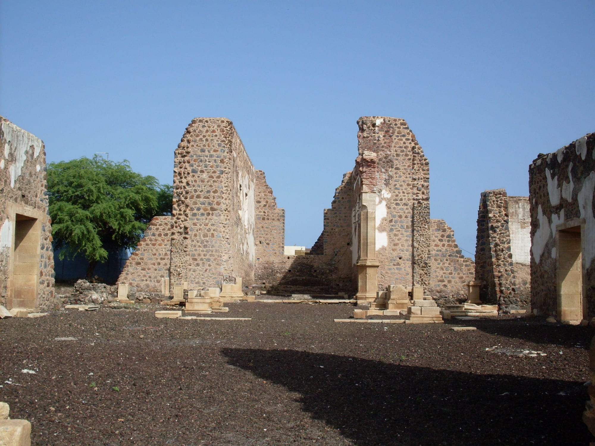 The roofless ruins of a large stone church, with tall fragments of rubble walls and pale cut-stone column bases on bare dark ground