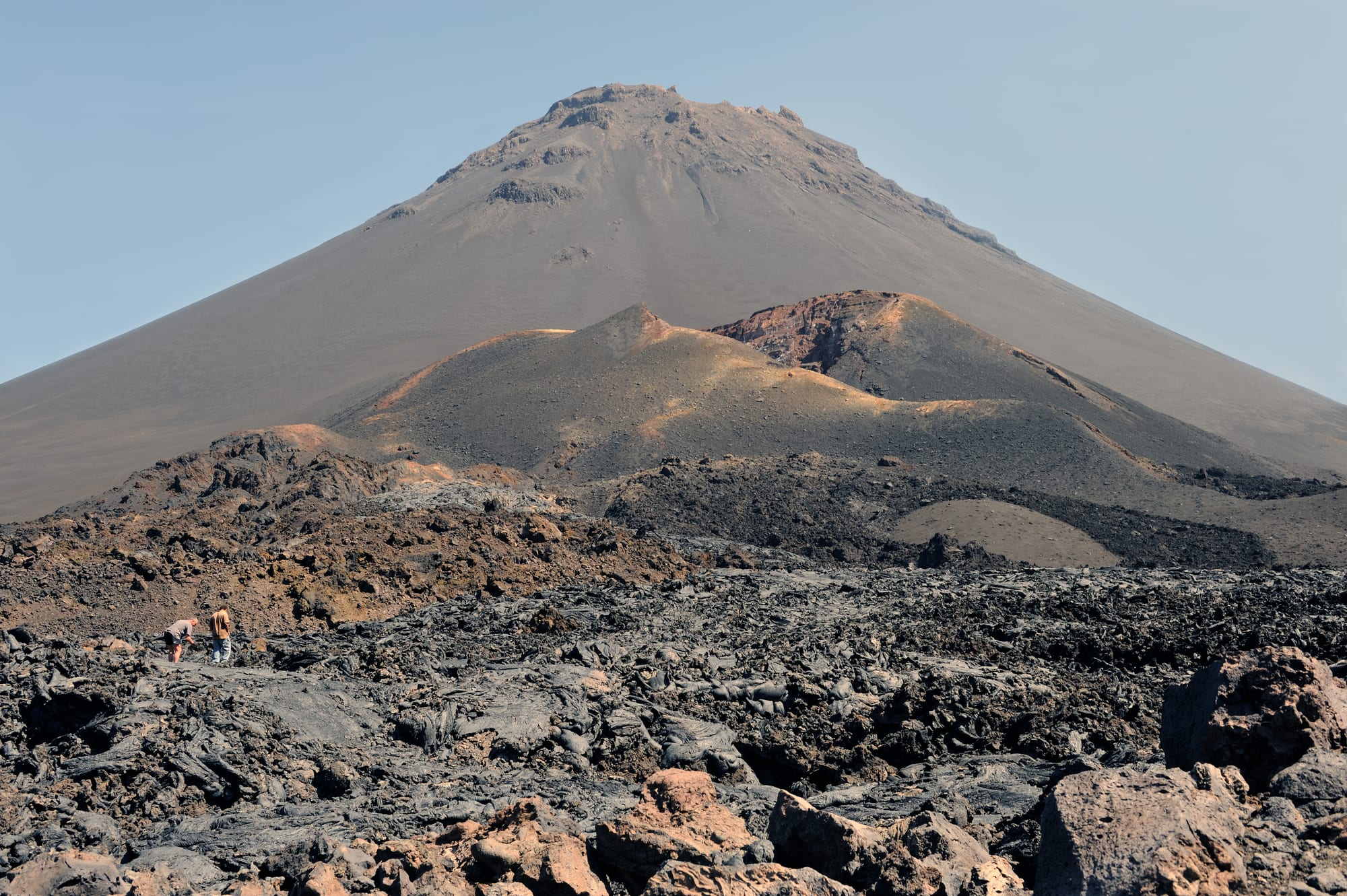 A large black volcanic cone rising from a caldera floor of dark lava and scattered white houses