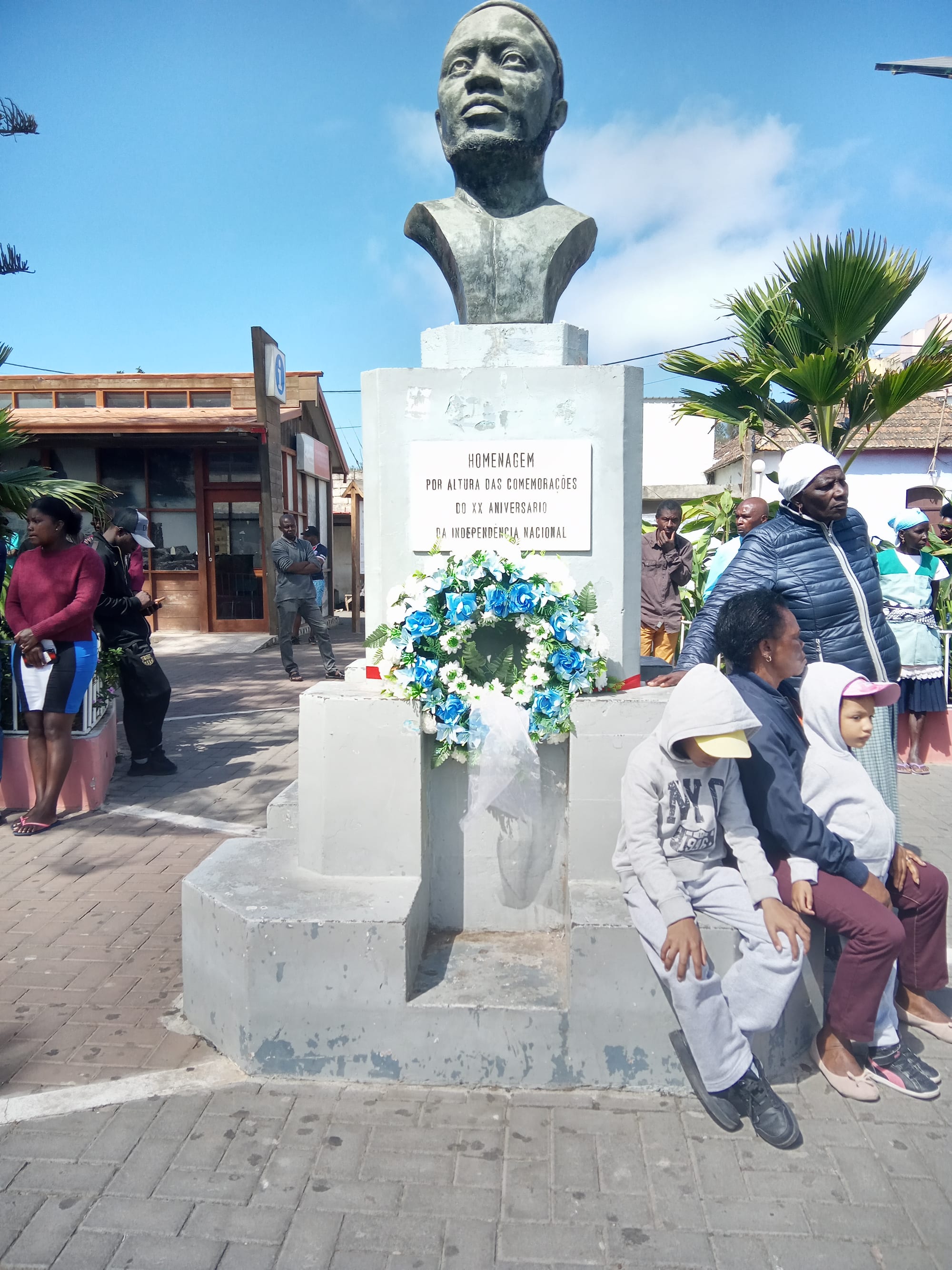 A weathered bronze bust of Amílcar Cabral in his knitted cap on a tall grey plinth in a paved square, with a wreath of blue and white flowers at its base and people sitting and standing nearby