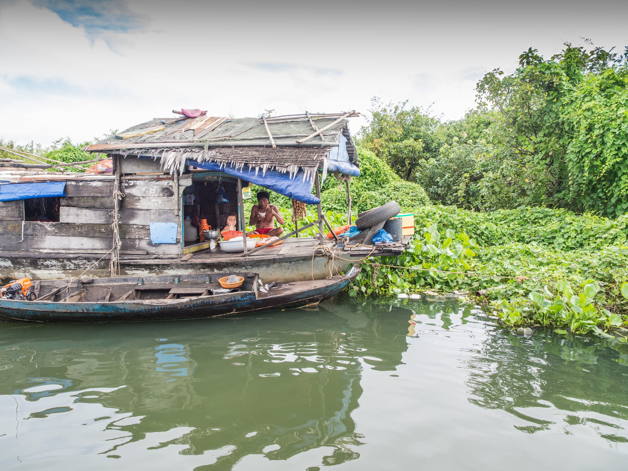 A father and child eat a meal together on the open deck of a weathered wooden floating house, moored among water hyacinth on the Tonle Sap