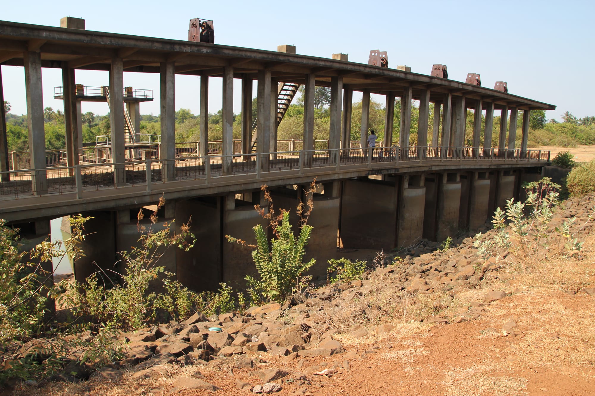 A long derelict concrete sluice structure on rows of pillars above a dry, rocky channel, with scrub growing around it