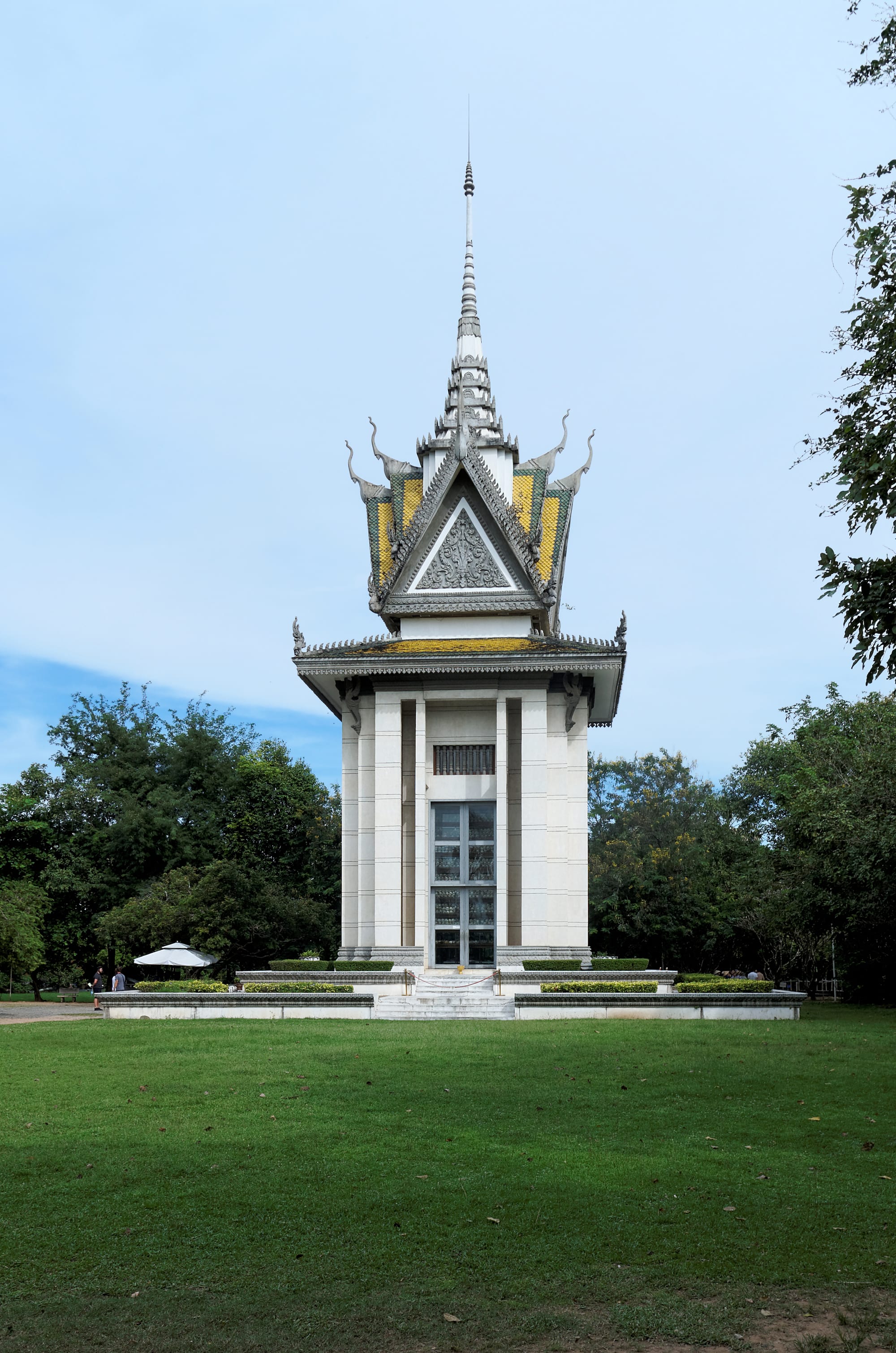 A tall white memorial stupa with glass sides filled with human skulls, on a quiet green site