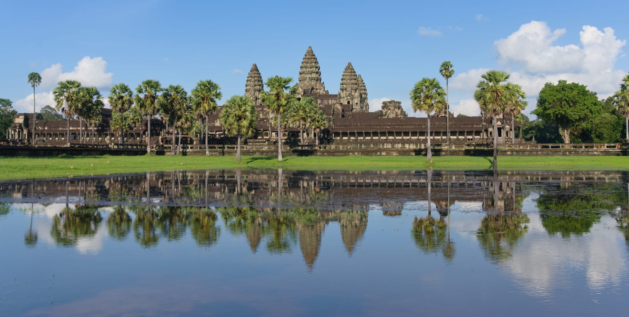 A vast stone temple with five lotus-bud towers mirrored in a still pond, framed by palms at dawn