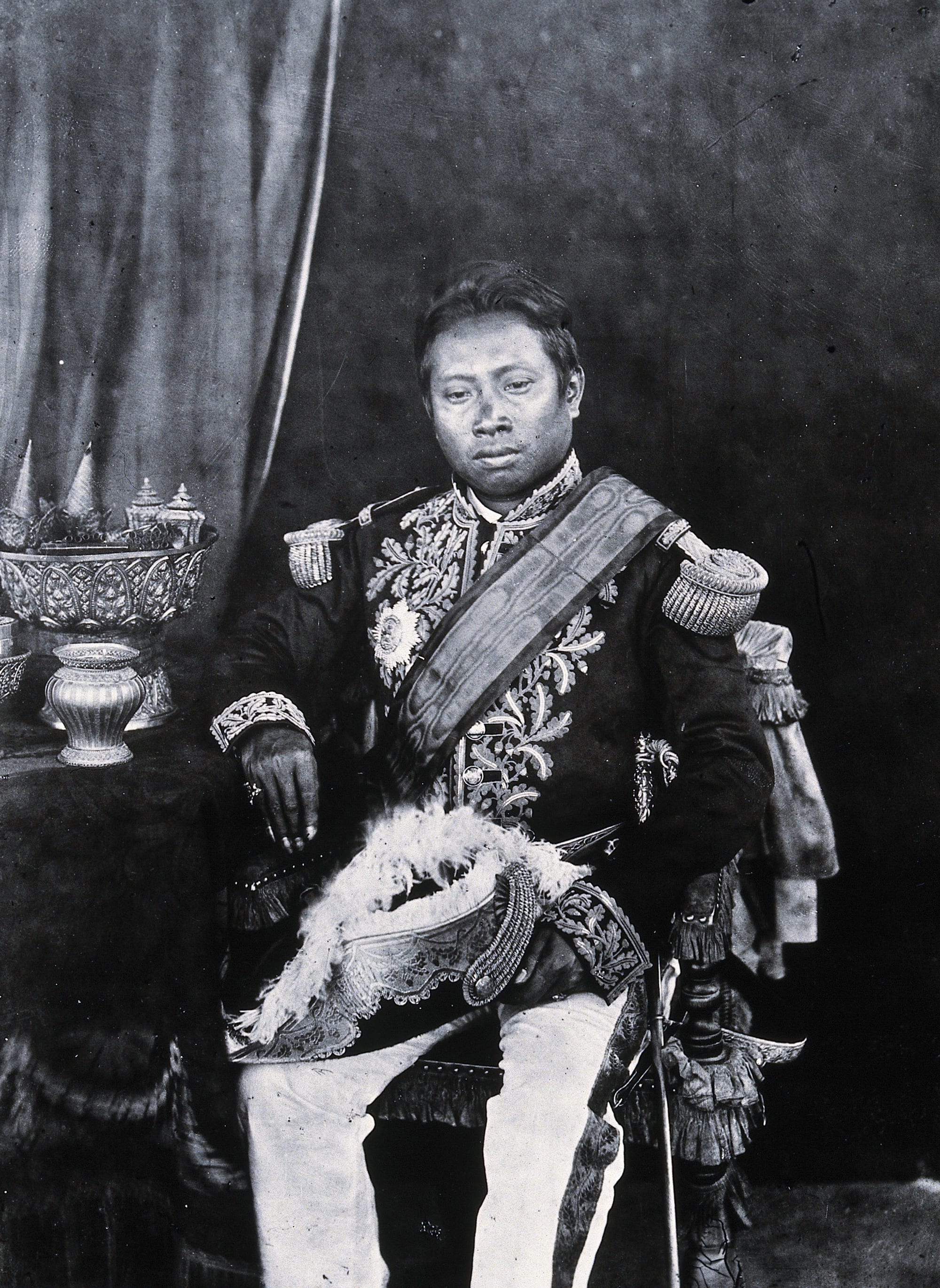 A 19th-century studio portrait of a seated king in a European-style embroidered dress uniform with epaulettes and a sash, a plumed hat on his knee and silver vessels on a table beside him
