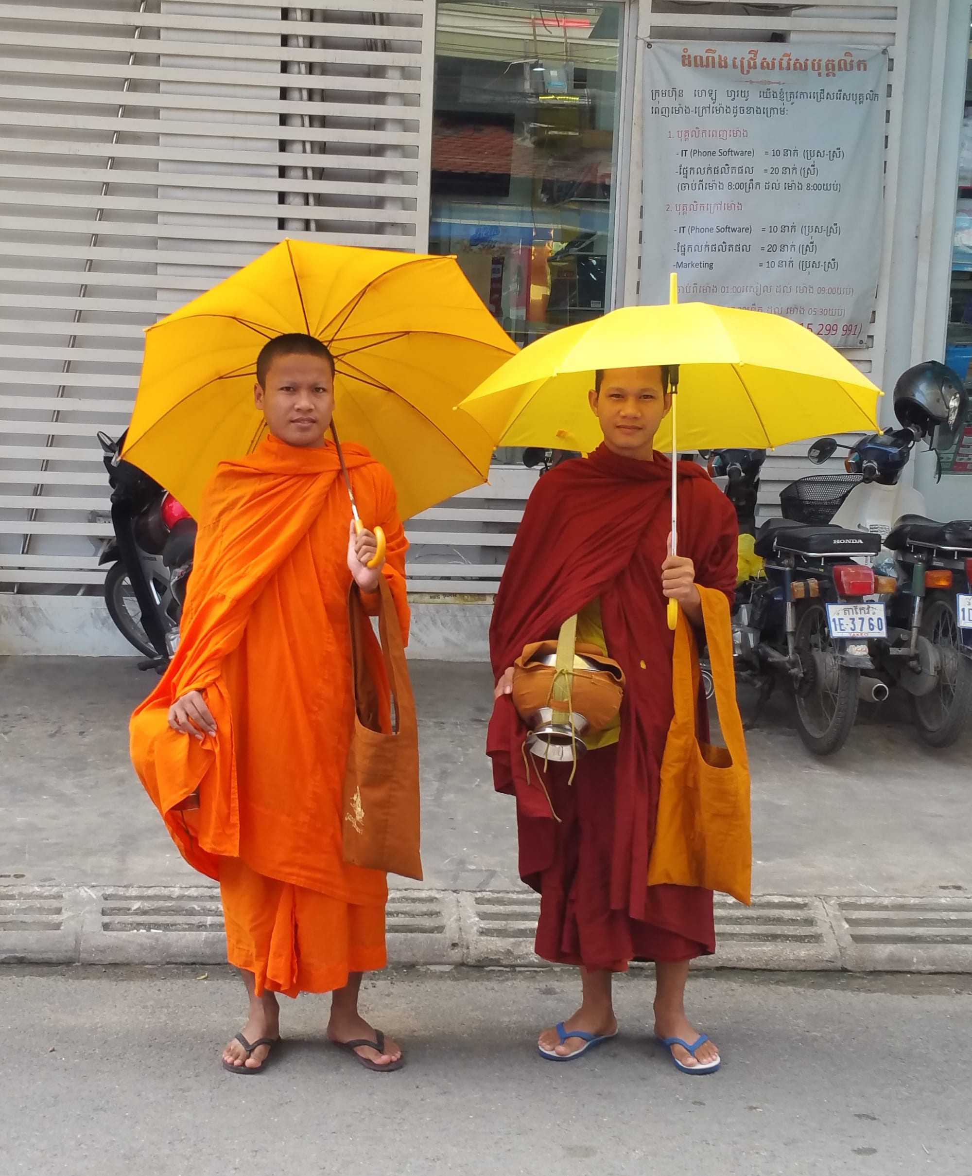 Two young Buddhist monks, one in bright orange robes and one in deep red, stand on a pavement holding yellow umbrellas, with an alms bowl and cloth shoulder bags