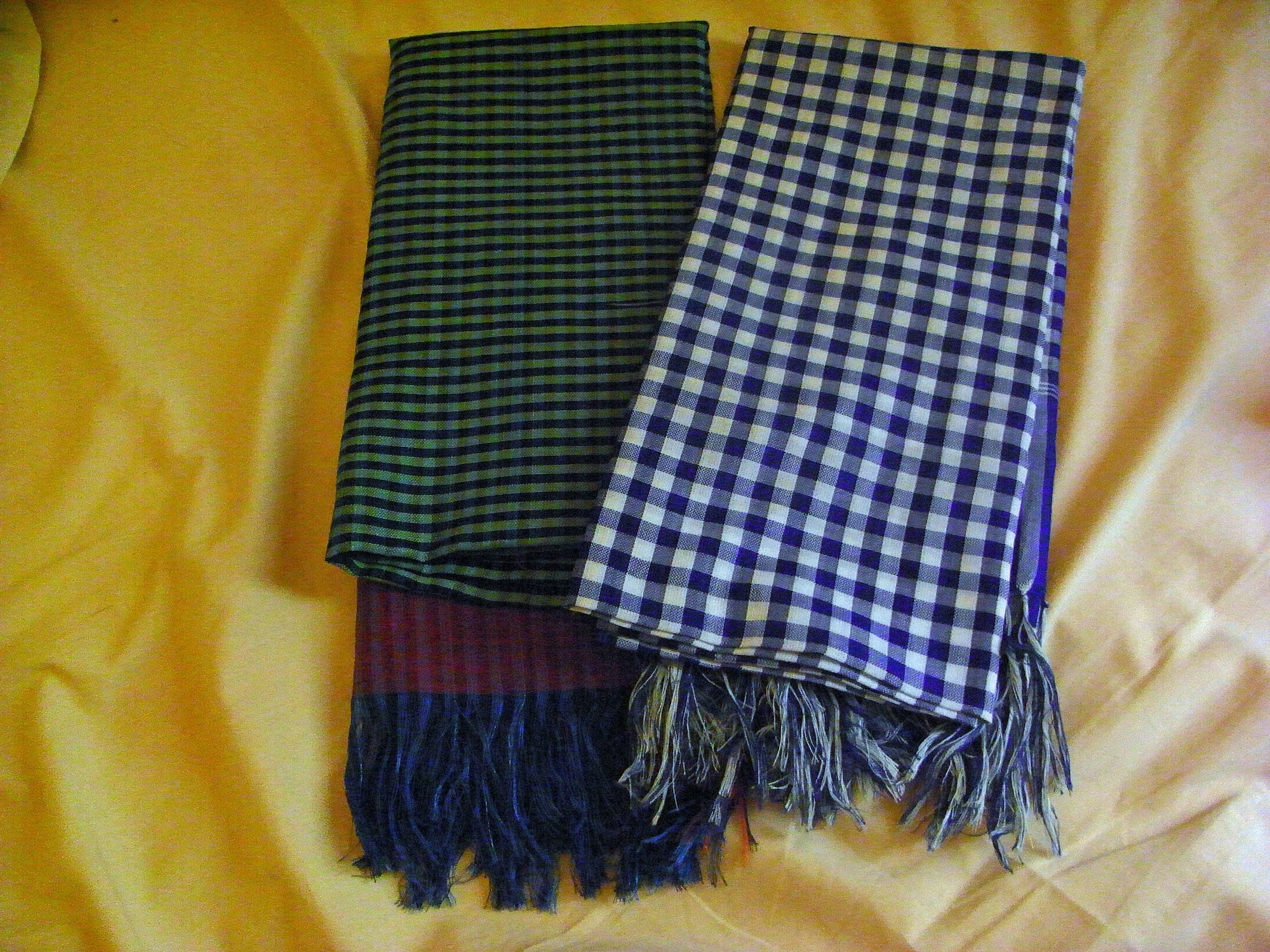 Two folded, fringed woven cloths, one in green and black stripes and one in a blue and white check, laid on a yellow fabric background