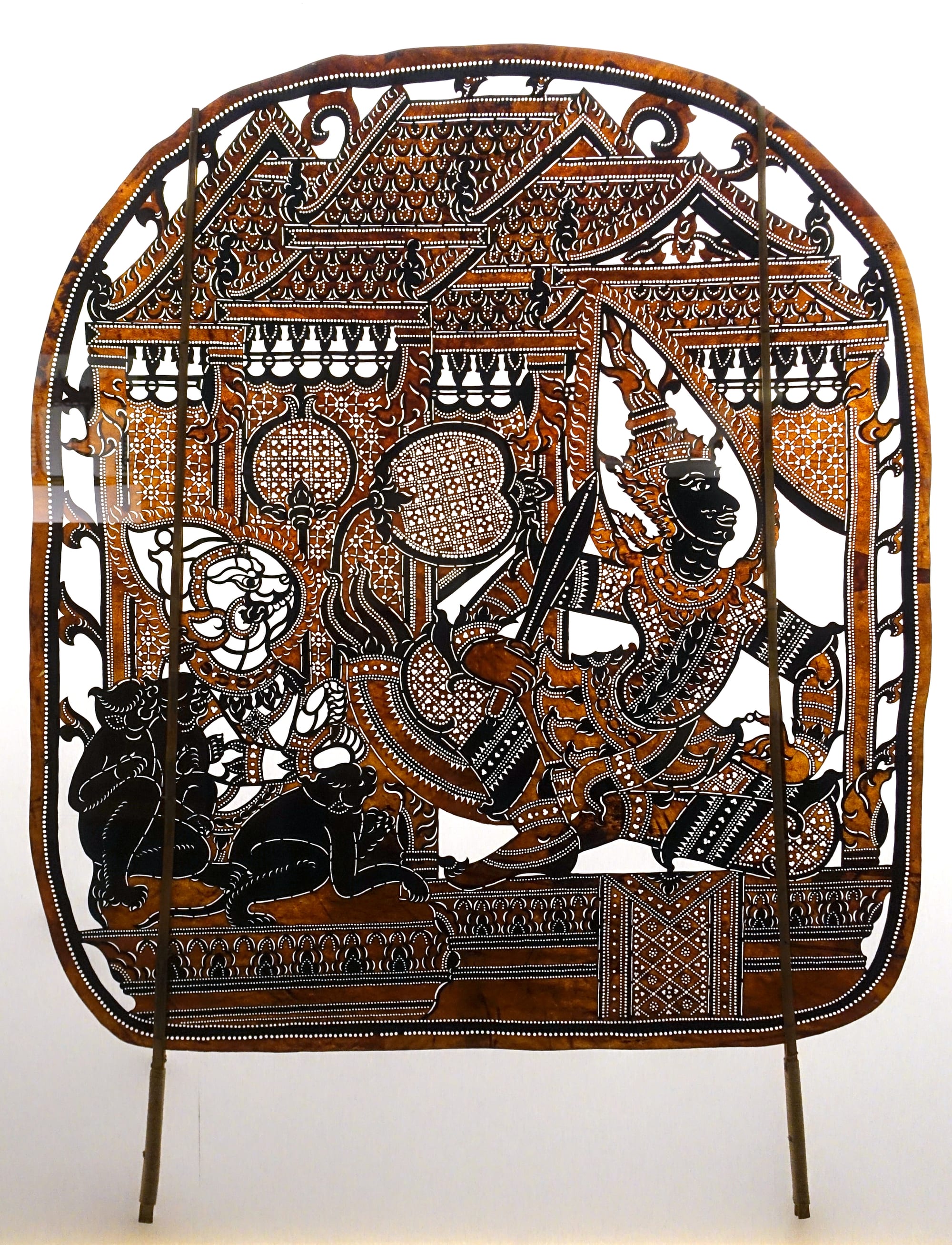A large leather shadow puppet cut in intricate openwork, showing a crowned hero in combat before an ornate palace, mounted on two bamboo sticks