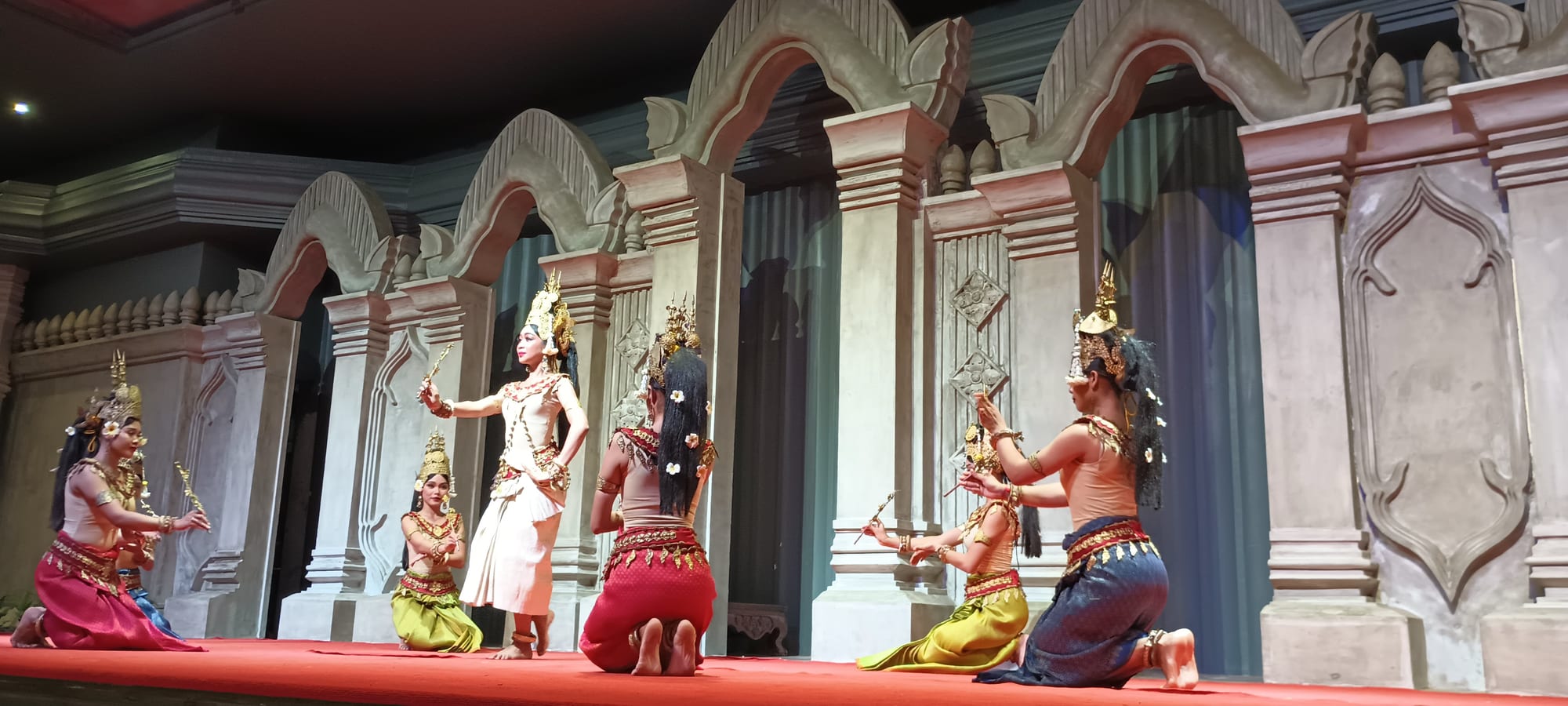Six dancers in gilded pointed crowns and brightly coloured silk costumes perform on a stage in front of carved stone-style arches, some kneeling and some standing, their hands held in curved, precise gestures