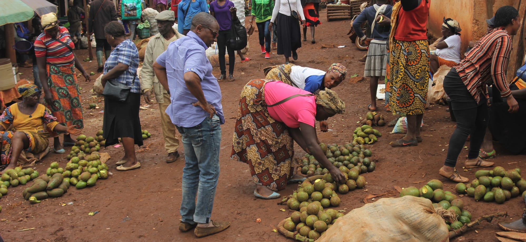 A crowded outdoor market with sellers and buyers gathered around large piles of avocados on the ground
