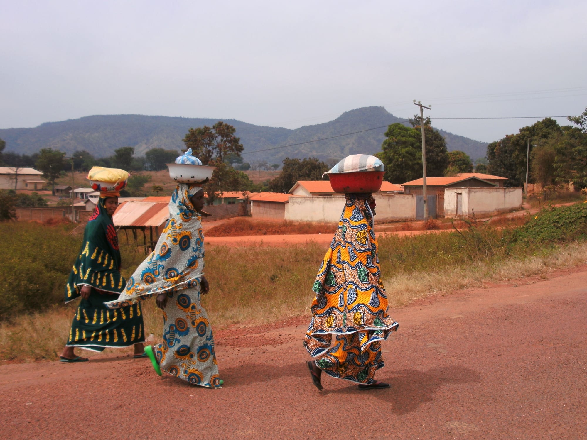 Three women in colourful patterned dress walking along a red dirt road, each carrying a bowl or bundle on her head, with houses and hills behind