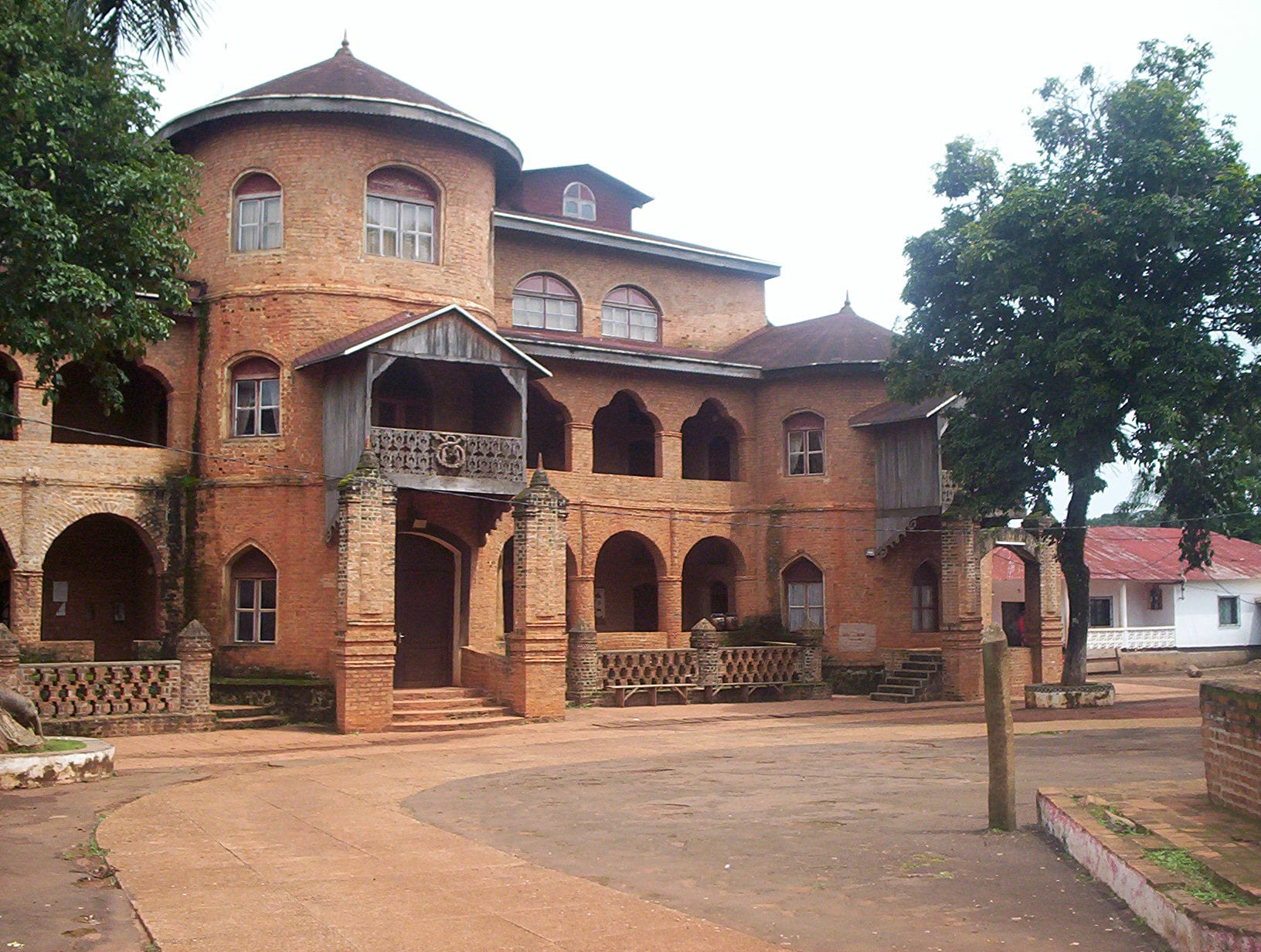 A large symmetrical two-storey palace with arched galleries and pointed roof turrets, in a dusty square