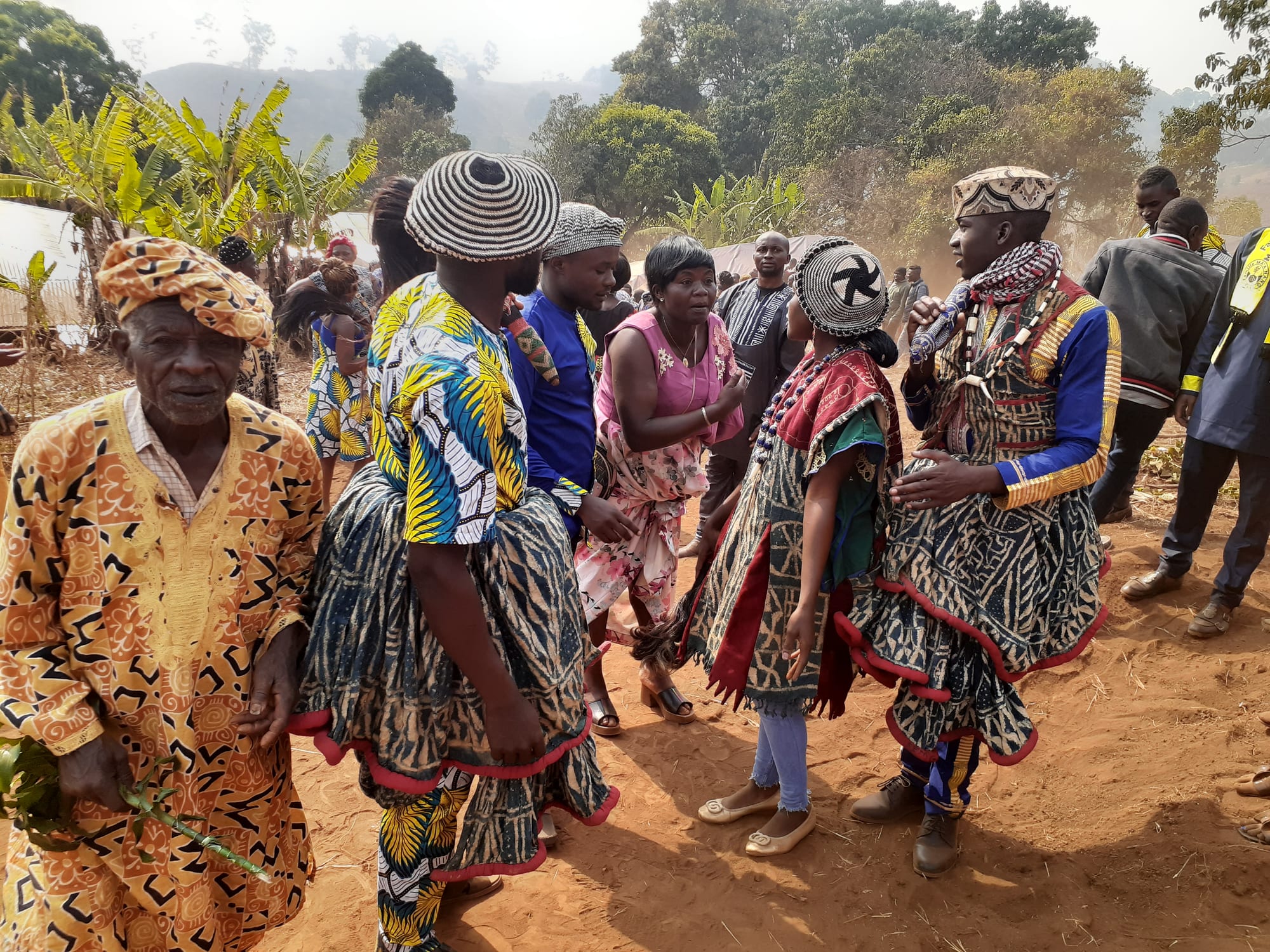 A crowd on a red-earth clearing, several people wearing indigo-and-white patterned robes with red trim and striped woven caps, an older man in an orange printed shirt in the foreground