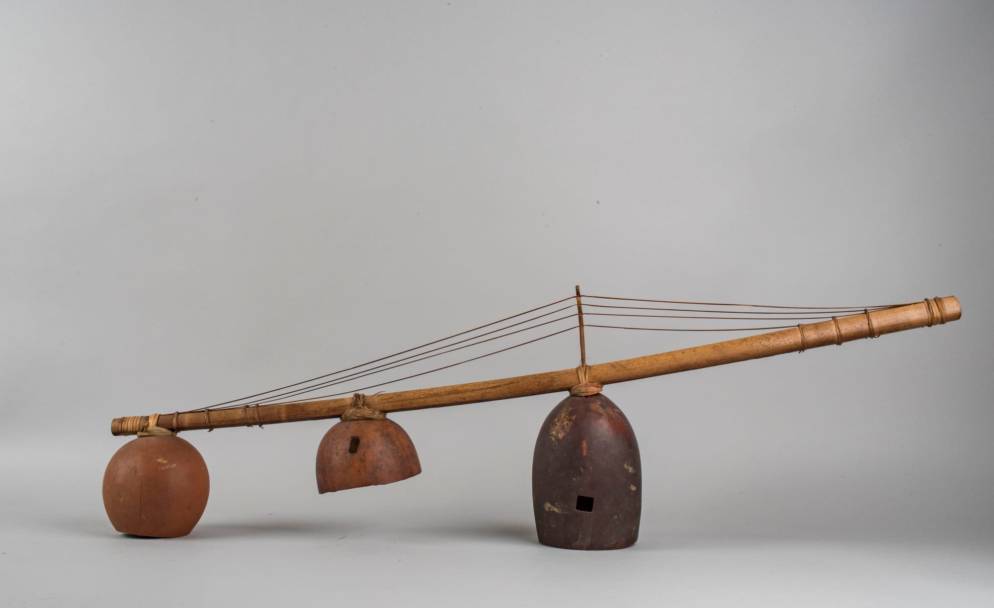 A long wooden stick with several fine strings raised on a central bridge, three hollow gourd resonators hanging beneath it, photographed against a grey background