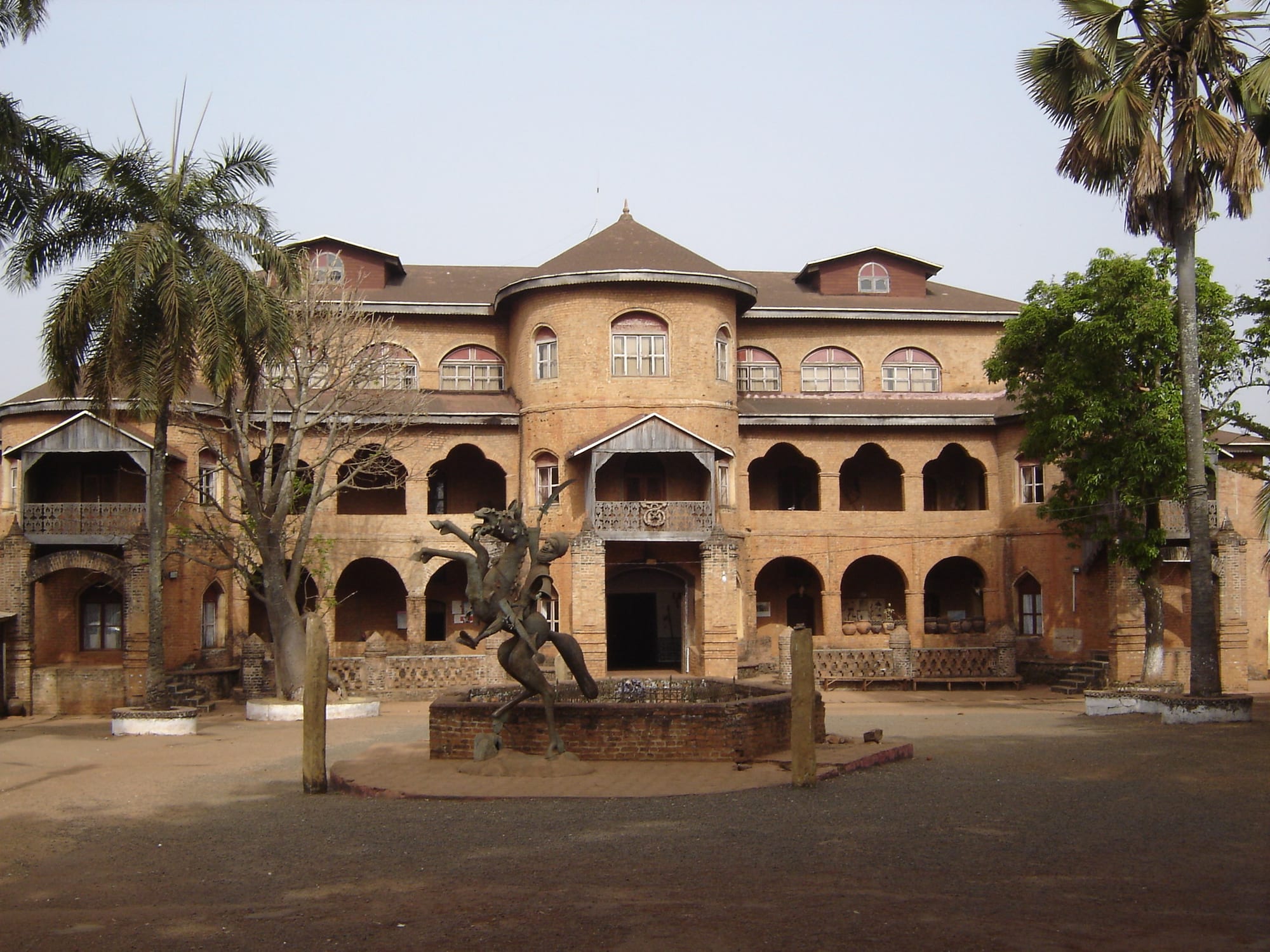 A large two-storey brick palace with arched verandahs, a central round tower and a bronze horse-and-rider sculpture on a plinth in the forecourt, framed by palm trees