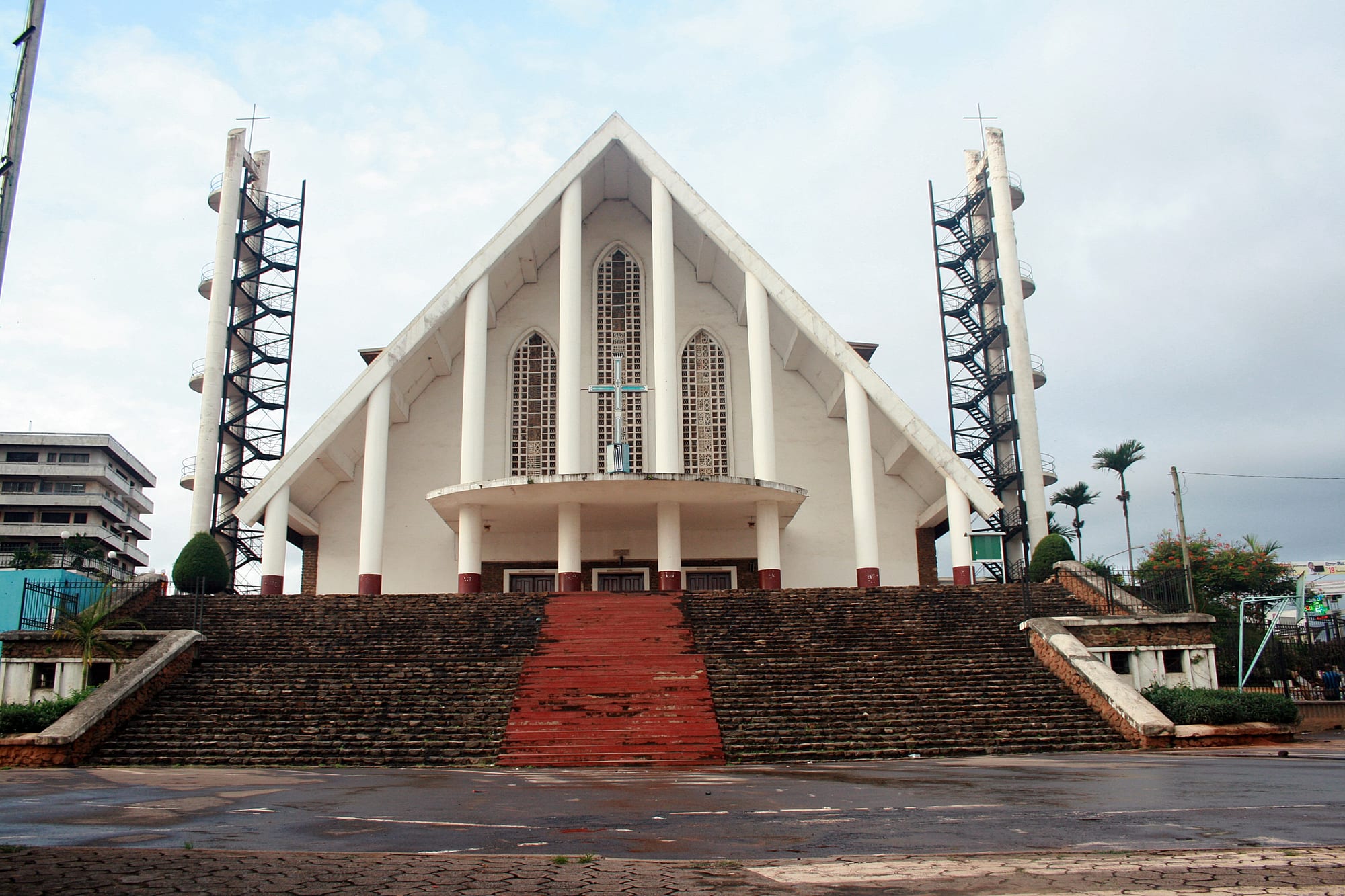 A large white cathedral with a steep triangular gable, tall pillars, arched windows and a cross, reached by a broad flight of stone steps with a red central stairway