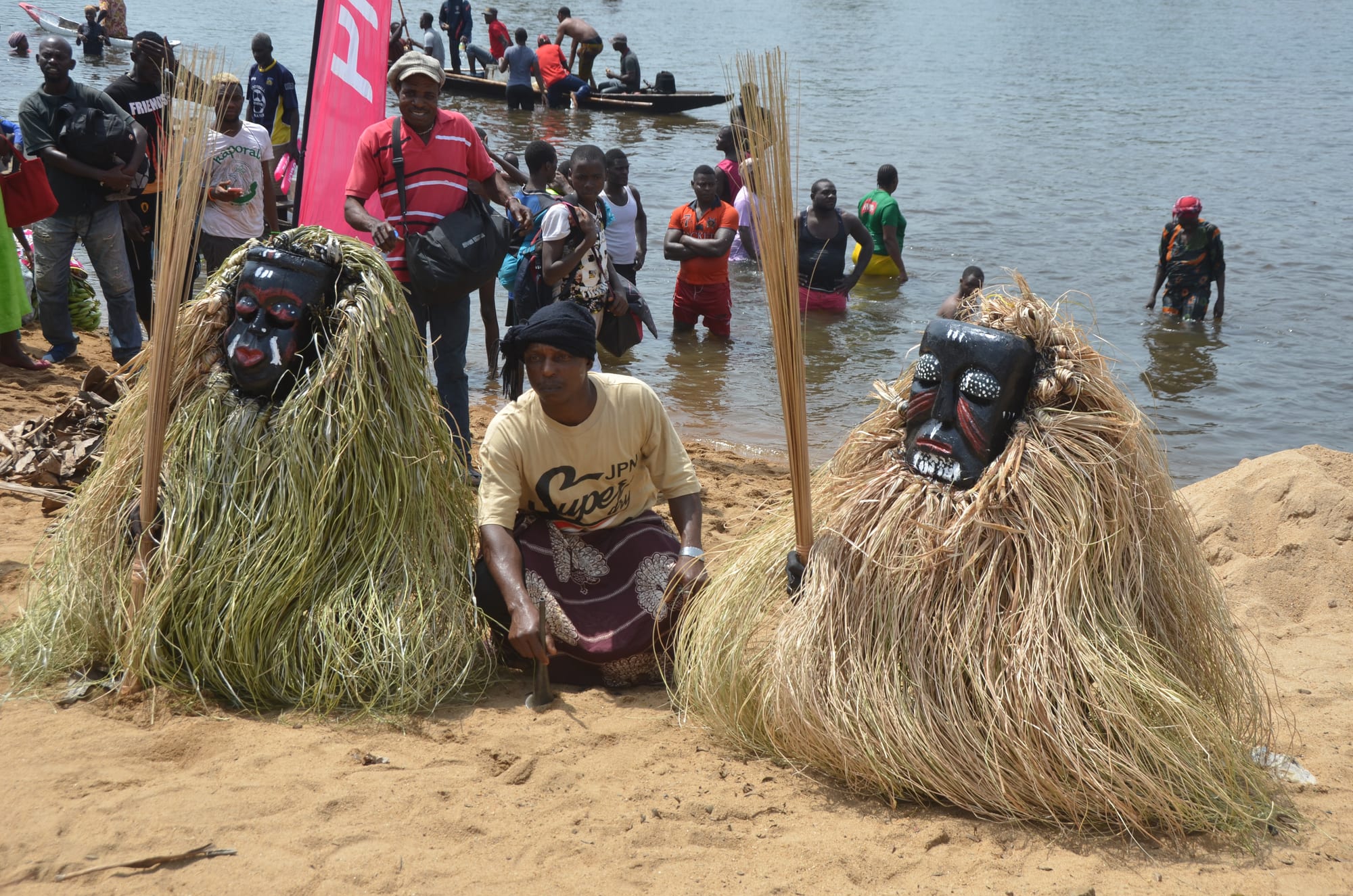 Two masked figures covered head to foot in long straw costumes, with carved dark wooden faces, sit on a sandy riverbank beside a woman in a cream T-shirt, with people wading in the river and canoes behind