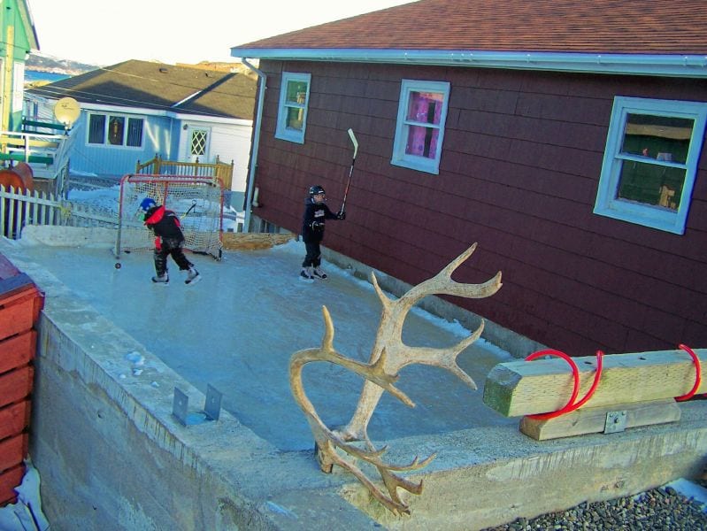 Two children playing hockey on a small backyard ice rink between suburban houses