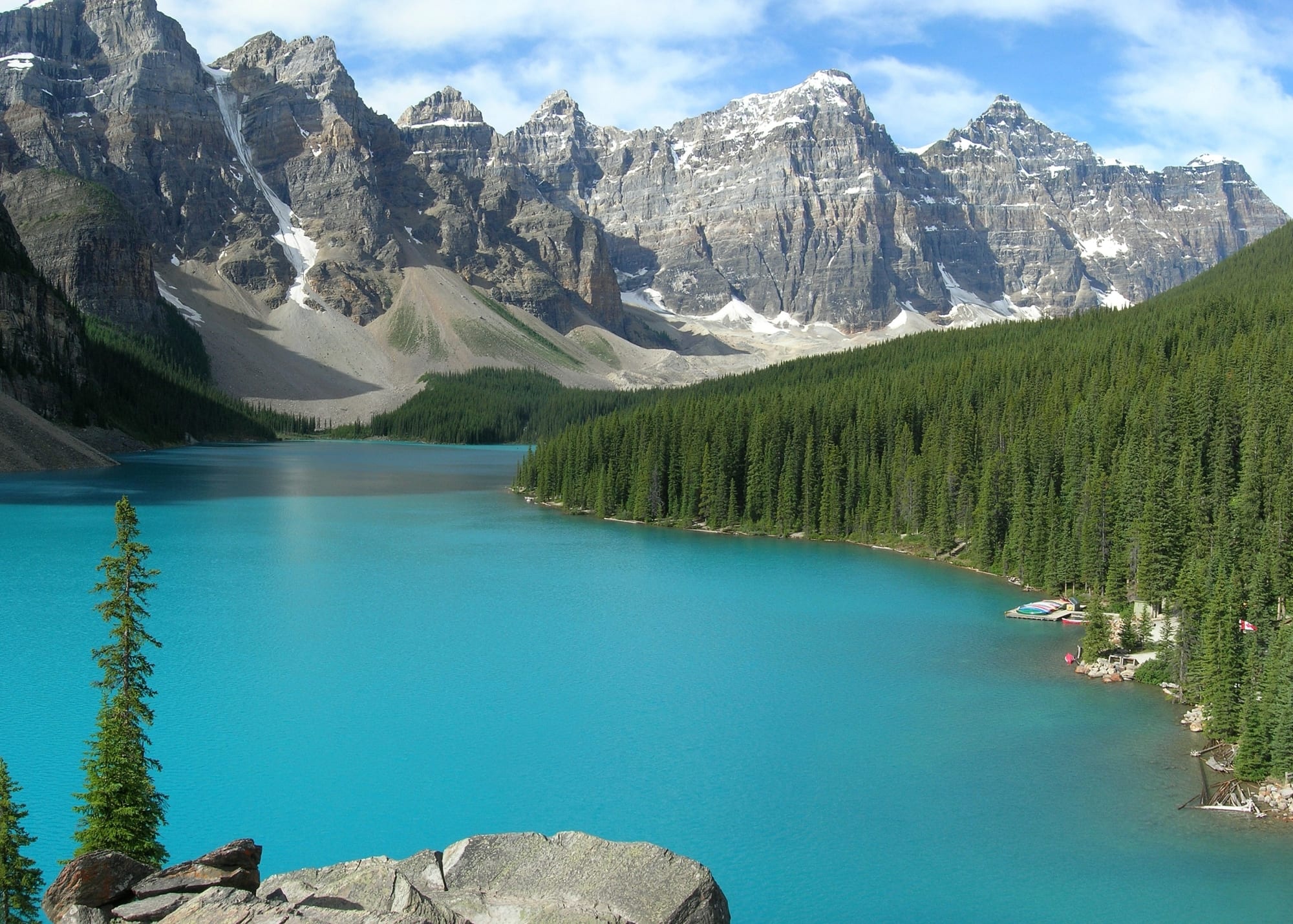 A brilliant turquoise lake below a row of sharp snow-streaked peaks, pine forest around the shore