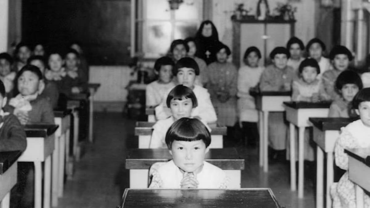 Rows of Indigenous children at wooden desks in a bare classroom with a nun and a priest, 1939