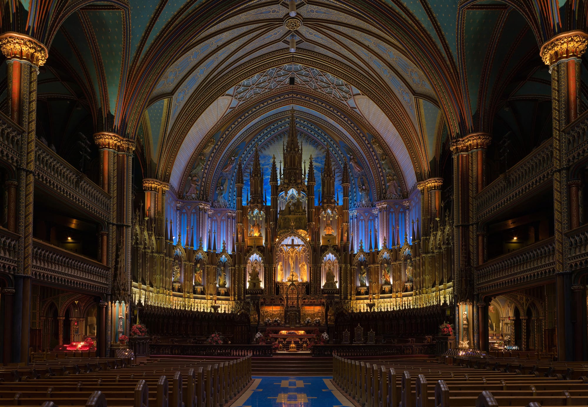 The nave of a Gothic Revival basilica, with rows of wooden pews leading to an ornate gilded altarpiece lit in deep blue, beneath a painted vaulted ceiling