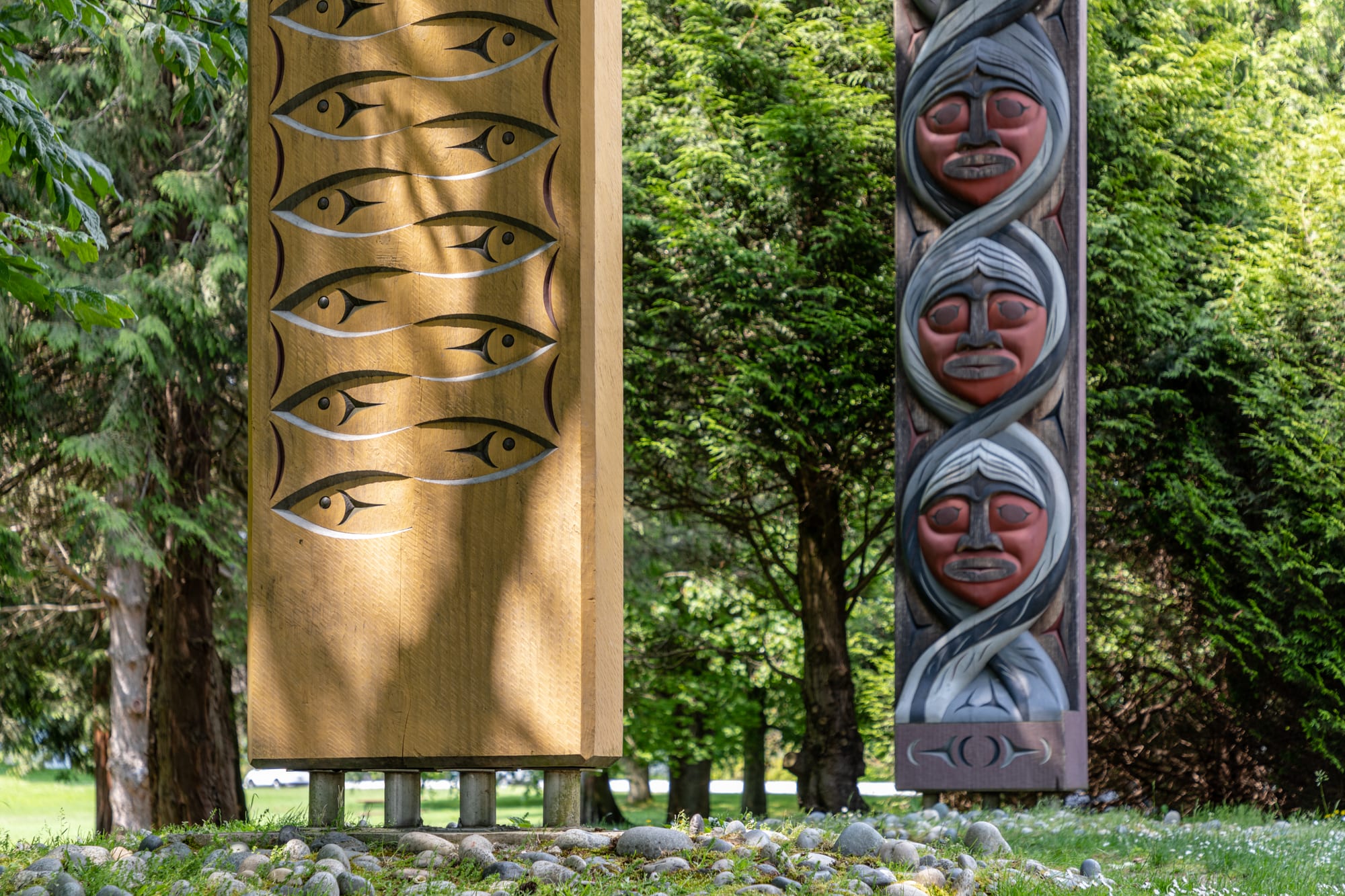 Two carved wooden posts among trees, one of pale wood carved with rows of stylised fish, the other a dark pole carved with three stacked red faces wrapped in grey bands