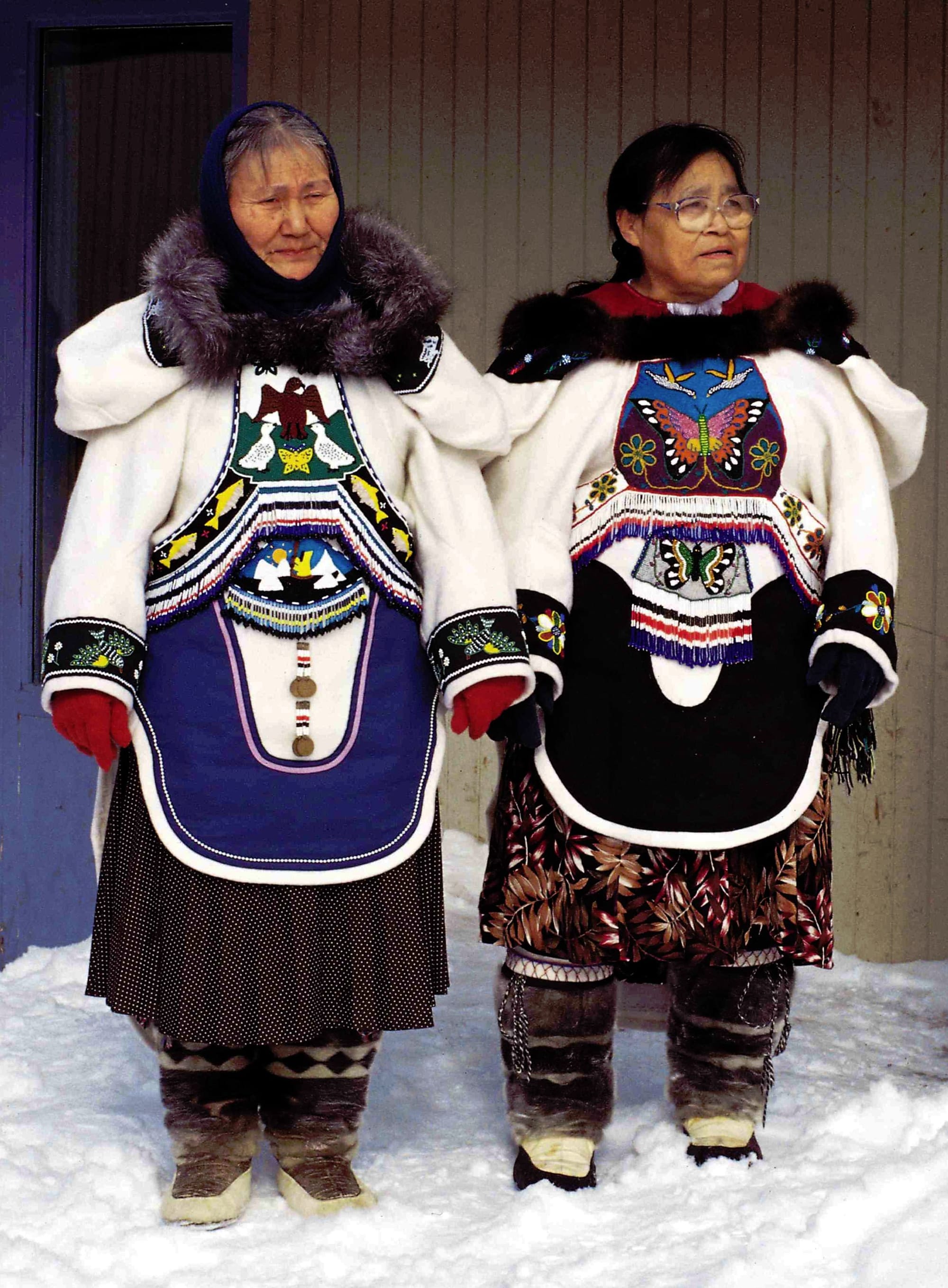 Two older Inuit women standing in snow, wearing white parkas with fur-trimmed hoods and elaborate beaded and embroidered panels showing birds and butterflies