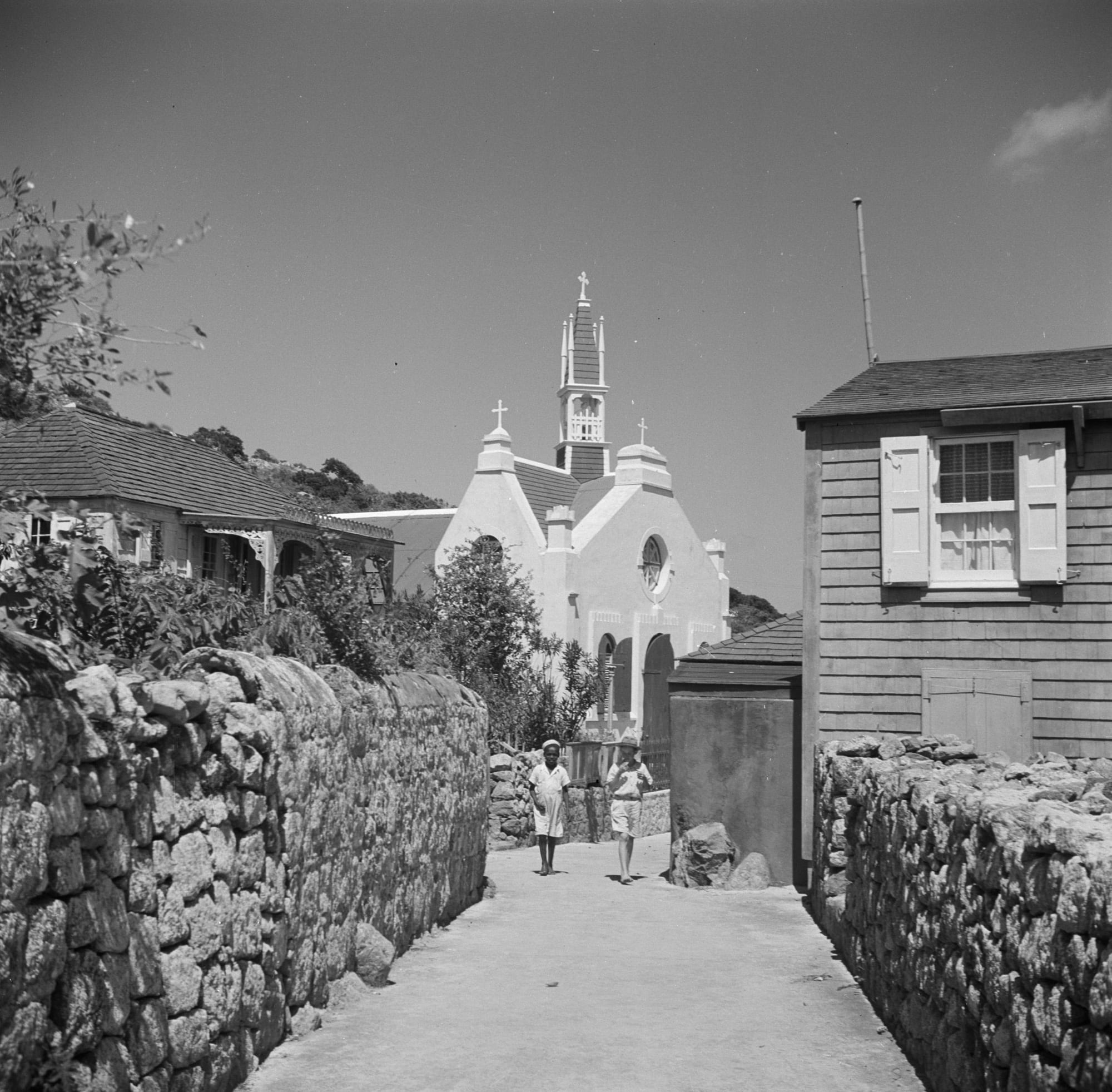 A black-and-white photograph of a narrow lane between dry-stone walls leading to a white church with a pointed bell tower and crosses, with a shingled house on the right and two people walking in the distance